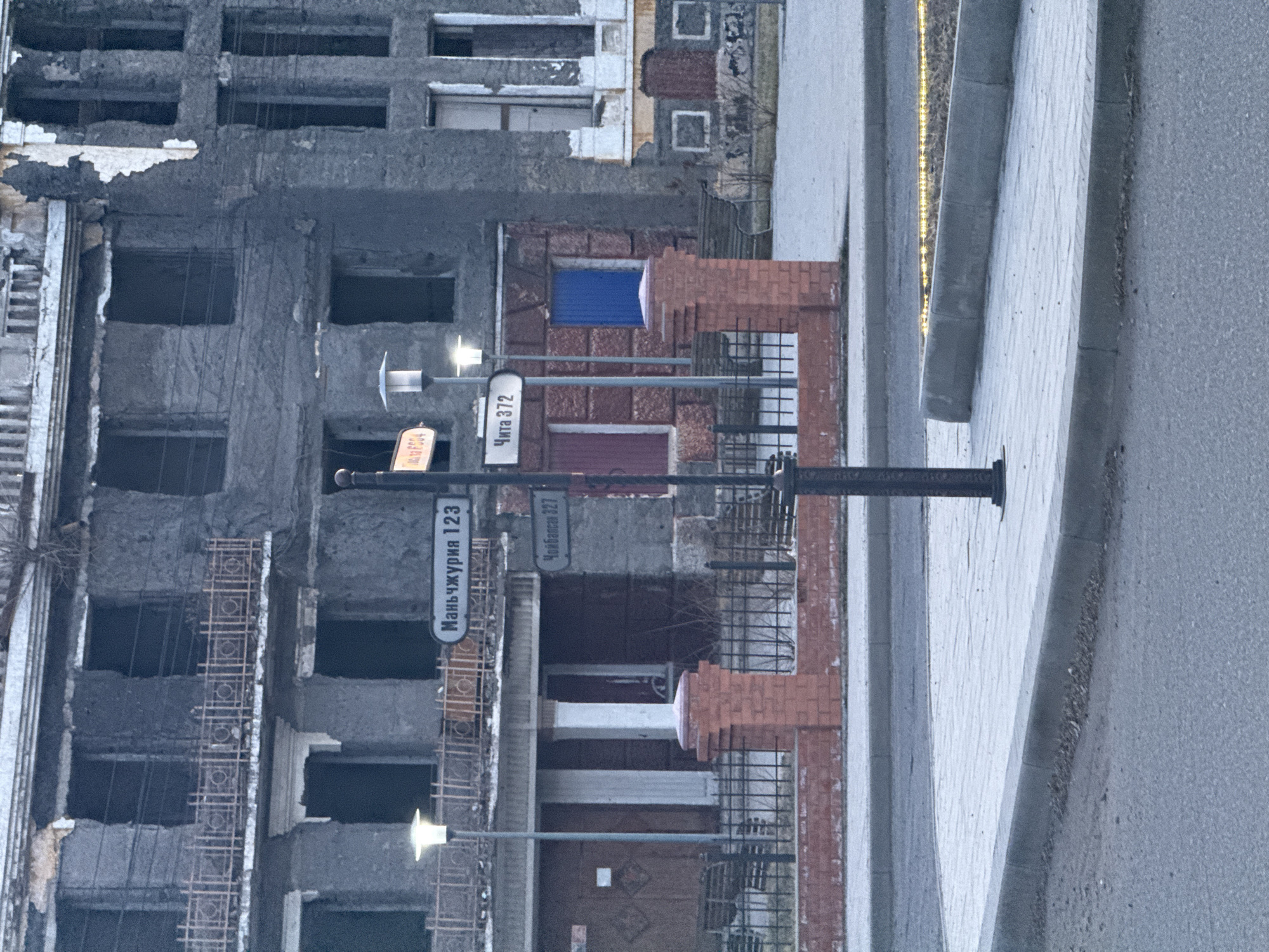 The image shows a street scene in Borzya, Russia. A severely damaged building stands adjacent to the sidewalk. The building has multiple window openings, many without glass. There is exposed rebar along the sides of the building. A street sign indicates "Маньчжурия 123" (Manchuria 123) and "Чита 372" (Chita 372), likely street names and numbers. Street lamps are present along the sidewalk. The sidewalk appears to be paved. Beyond the sidewalk, a road is visible. A brick building is partially visible between the street sign and the building.
