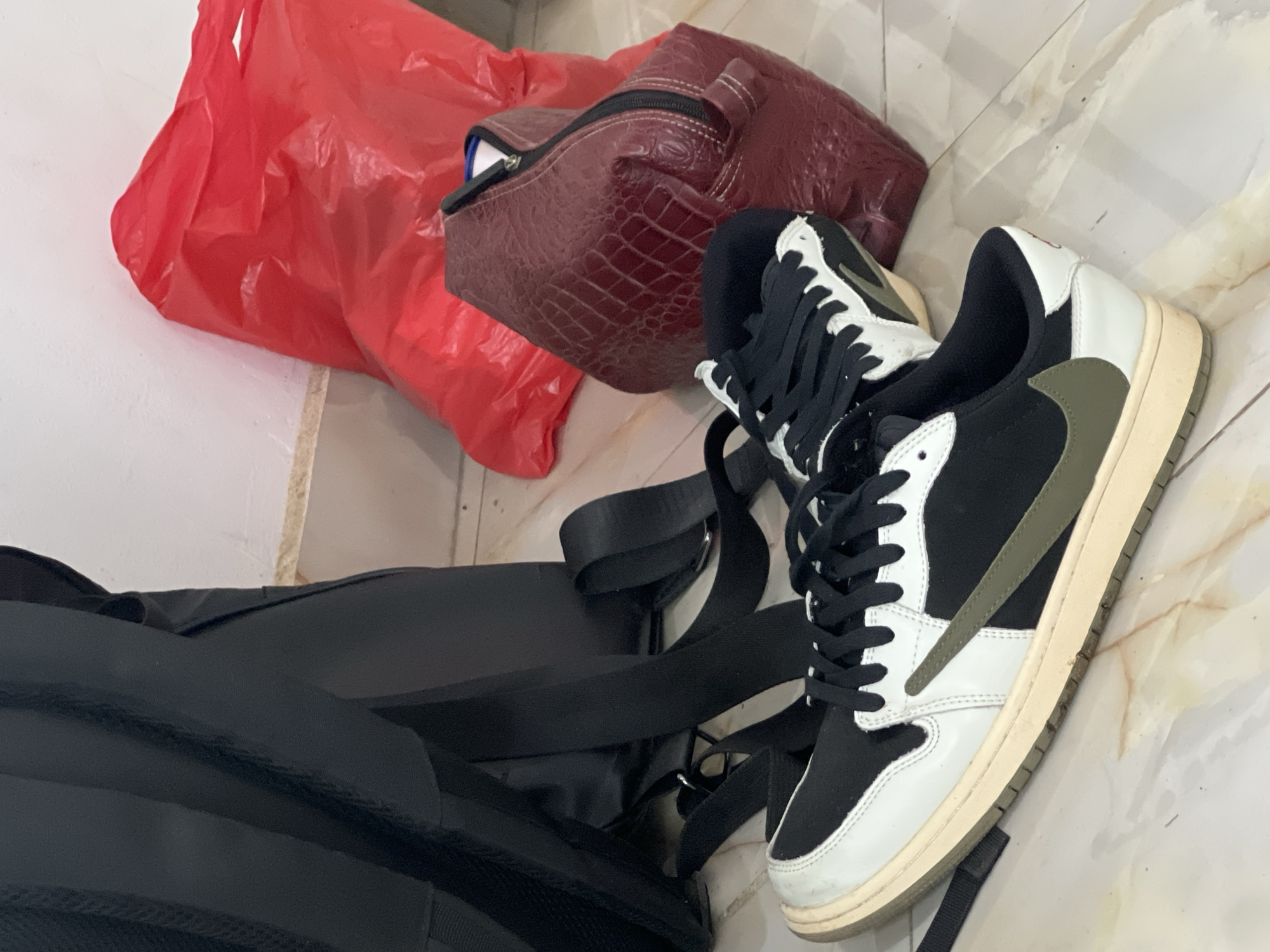 This image captures an indoor scene in Abuja, Nigeria, featuring various personal items laid out on a light-colored, possibly beige or white, tiled floor next to a plain, light-colored wall.
On the left, a crumpled red plastic bag occupies a significant portion of the frame. Behind it, towards the center, rests a small, dark red or maroon bag or pouch. This bag exhibits a distinct textured surface, resembling crocodile skin, and is secured with a black zipper closure.
In the bottom left and extending towards the center, a black backpack or large bag is partially visible, with its black fabric straps unfurling across the floor.
Dominating the right side of the image is a pair of high-top sneakers. These shoes are primarily black and white, featuring black laces and a notable military green (or olive green) Nike swoosh on the sides. The soles of the sneakers are a cream or off-white color and show slight signs of wear.
There is no specific activity depicted; the items appear to be in a temporary state, perhaps packed, unpacked, or simply stored. No individuals are present. The setting suggests a casual, possibly domestic environment. The time of day and weather conditions are not discernible from the image. No visible text can be identified on any of the objects.