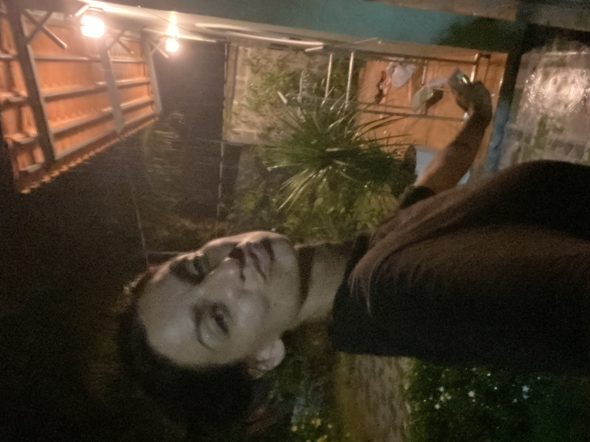 The image captures a woman taking a selfie at night in an outdoor setting. She is wearing a dark t-shirt and has her hair pulled back. Her facial expression is one of pursed lips. The background features a tiled roof with overhead lighting, suggesting a covered outdoor area like a patio or veranda. To the right, a wall and some vegetation are visible. In the distance, there appears to be a metal railing and some items on the ground. The ground is wet, possibly from rain. The overall lighting suggests it is nighttime. The scene is likely located in Mũi Né, Vietnam, given the presence of tropical-looking plants and the style of the outdoor structure.