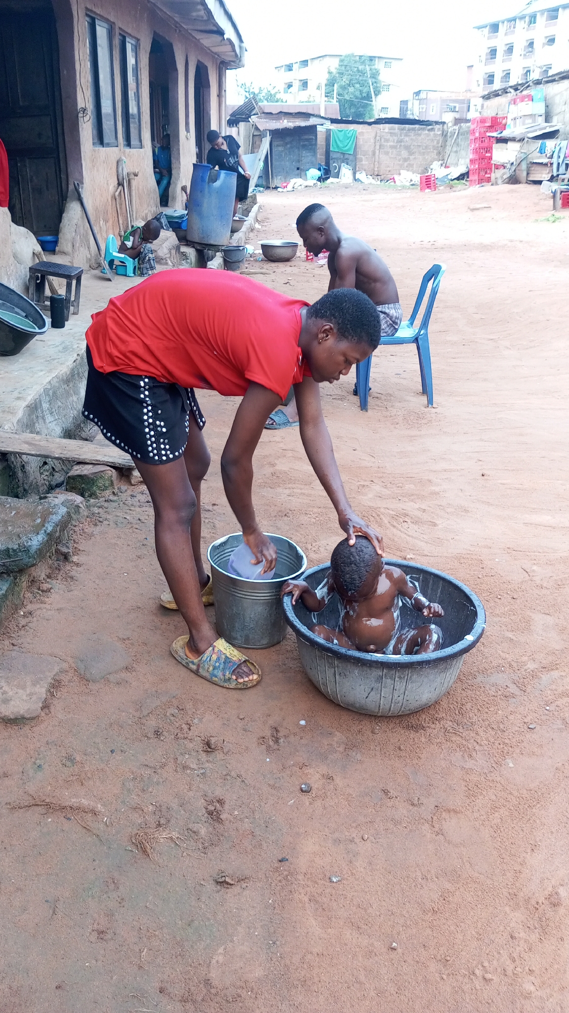 This candid daytime image portrays a daily life scene in Onitsha, Nigeria. The primary focus is a woman diligently bathing a small baby outdoors. The woman, dressed in a red t-shirt, black shorts with white embellishments, and patterned yellow sandals, leans over a large, dark plastic basin where the baby sits, partially submerged in water. She holds a small purple scoop in a metal bucket beside the basin, her right hand gently supporting the baby's head. The baby appears to be covered in soap or suds.
The ground is unpaved, consisting of reddish-brown dirt, with scattered small stones and debris. In the midground, to the left, a light brown, traditional-style building with arched doorways and rectangular windows stands. A blue plastic barrel and other containers are positioned near its base. Further back and to the right, a young man with short hair, shirtless, sits on a blue plastic chair, observing the scene or resting. Another person is partially visible near the building on the left.
The background reveals more of the surrounding environment: an expanse of dirt leading to more structures, including stacks of bright red plastic crates that suggest commercial or storage use. In the far distance, several multi-story buildings are visible under a clear or slightly hazy sky. The scene depicts a moment of nurturing care within a bustling outdoor residential or mixed-use area, characteristic of a warm climate. No visible text is present in the image.