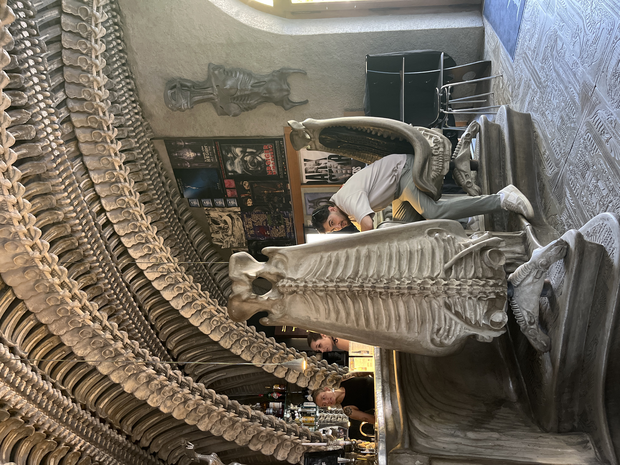 The scene is the interior of a bar designed with a skeletal, biomechanical aesthetic, strongly reminiscent of H.R. Giger's art. The ceiling is an arched structure resembling ribs and vertebrae, constructed from a light-colored material. A bar area is visible to the left, with bottles and glasses on shelves. Two women are behind the bar, one in a black tank top with tattoos on her arm, the other in a dark top. In the foreground, a large, sculpted skeletal structure resembling a ribcage with a skull-like head serves as a chair. A man in a light-colored t-shirt and gray pants is seated in this chair, looking towards the camera. The walls are decorated with posters, some featuring "H.R. Giger," and a sculpted bust of a biomechanical figure. The floor is intricately patterned with raised designs. The overall impression is that of a unique and immersive themed establishment.