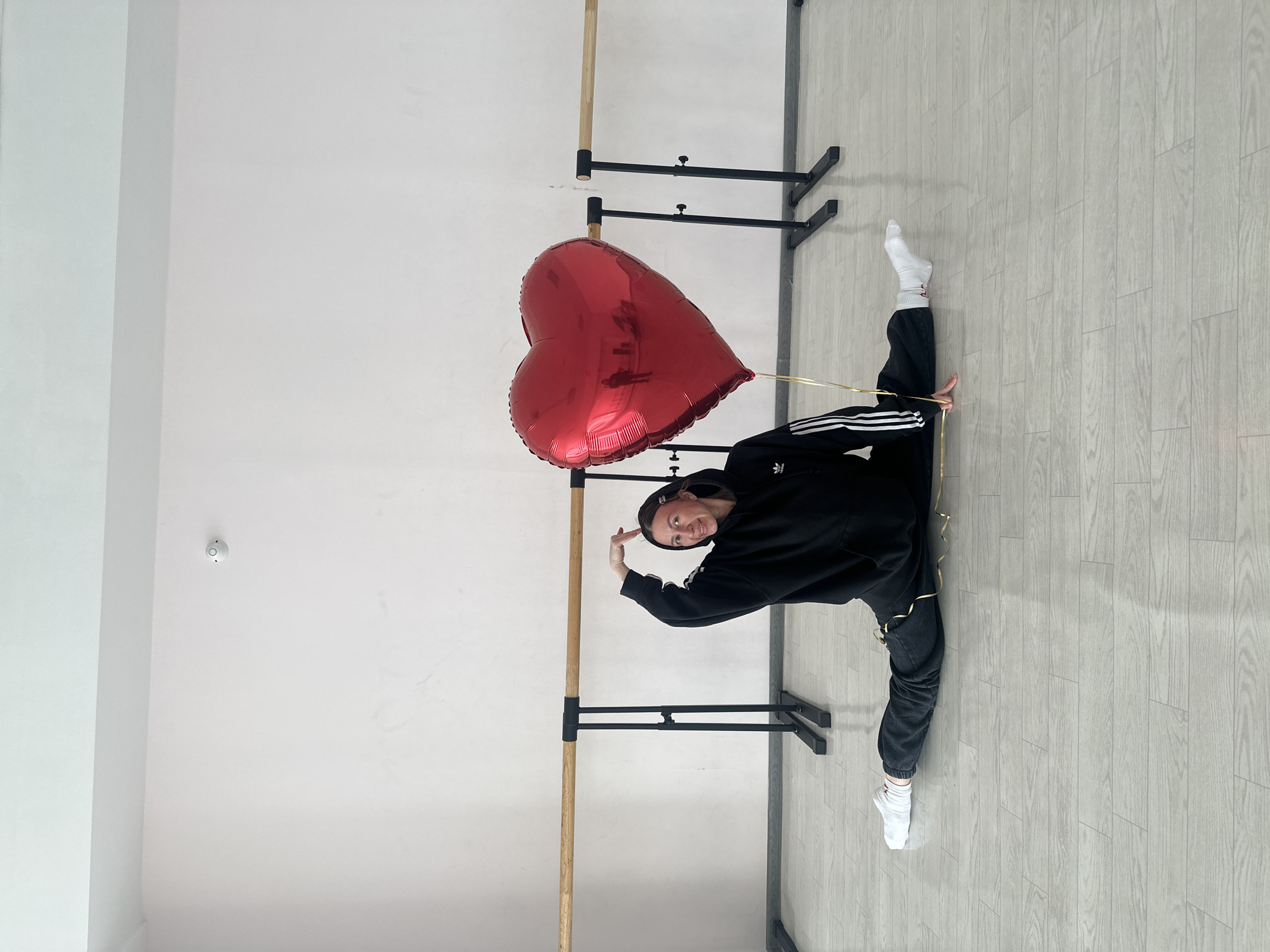 A person is performing a split while holding a large red heart-shaped balloon. The person is wearing a black Adidas tracksuit, a hooded sweatshirt, and white socks. The setting appears to be a dance studio with light-colored wood-look flooring, white walls, and a ballet barre mounted to the wall. The location is identified as Votkinsk, Russia.