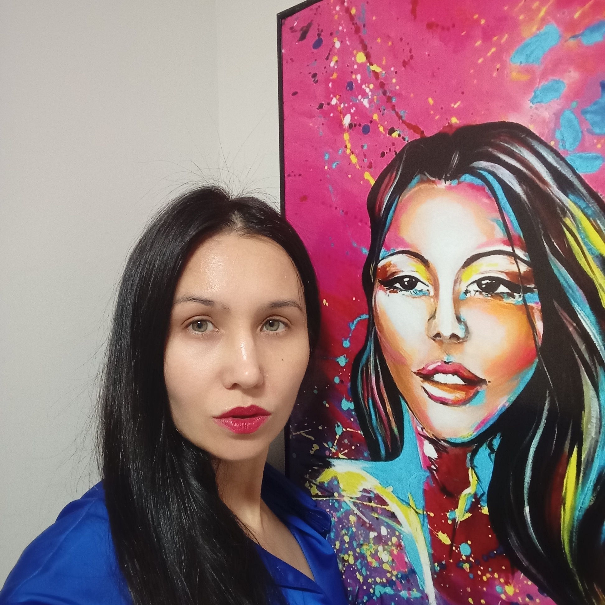 A woman stands next to a large portrait. The woman has long, straight black hair, fair skin, light-colored eyes, and is wearing red lipstick. She is wearing a blue garment. To her right is a framed portrait of a woman. The portrait features a woman with similar features to the real woman, with dark hair and a fair complexion. The portrait is brightly colored with pink, blue, yellow, and white paint. Splatters of paint decorate the background of the portrait. The background of the photograph is a plain white wall. The location is unknown, and information about Pervomaysky, Russia is not discernible from the image.