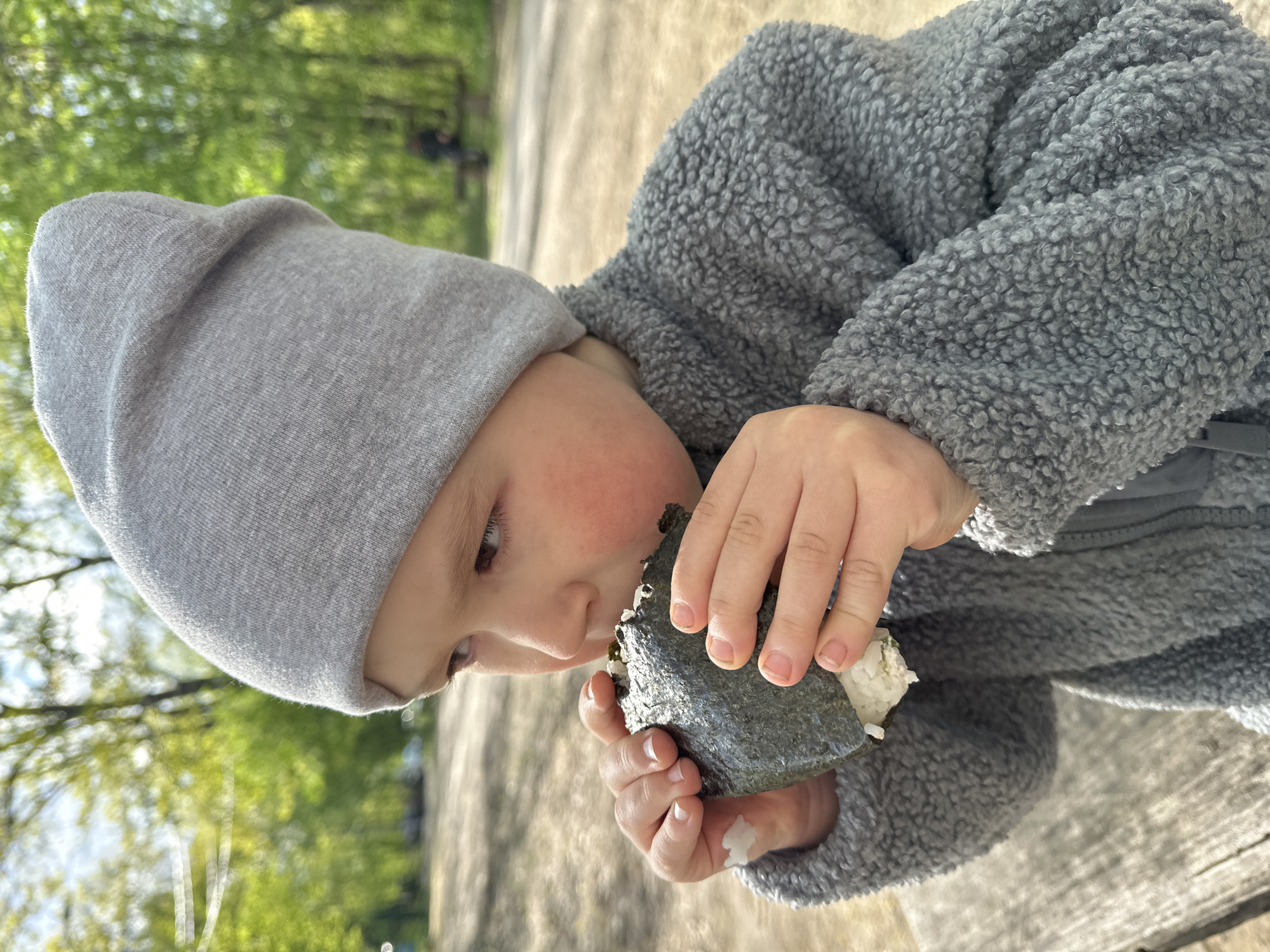 A child wearing a gray hat and a fleece jacket is eating onigiri. The child has rosy cheeks. The background is blurred, but trees with green leaves can be seen. The location could be a park or outdoor eating area in Nizhny Novgorod, Russia.