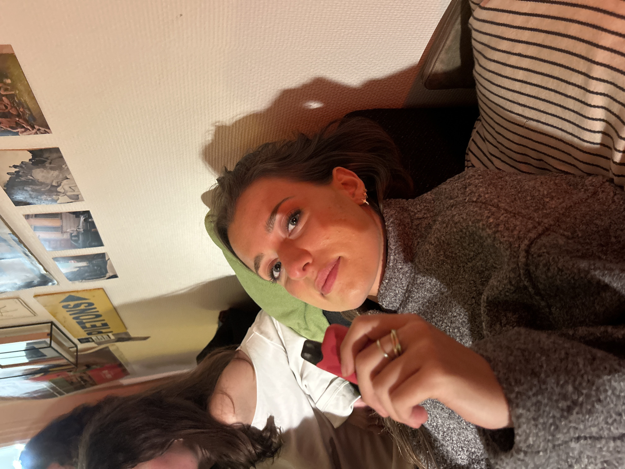A low-angle, medium shot depicts a young woman lying on a striped pillow and a green pillow. She is wearing a gray sweater and holding a red object. A second person is partially visible at the bottom of the frame, wearing a white shirt. Pictures and a sign reading "PIETONS" are mounted on the wall. The location is in an unknown city.