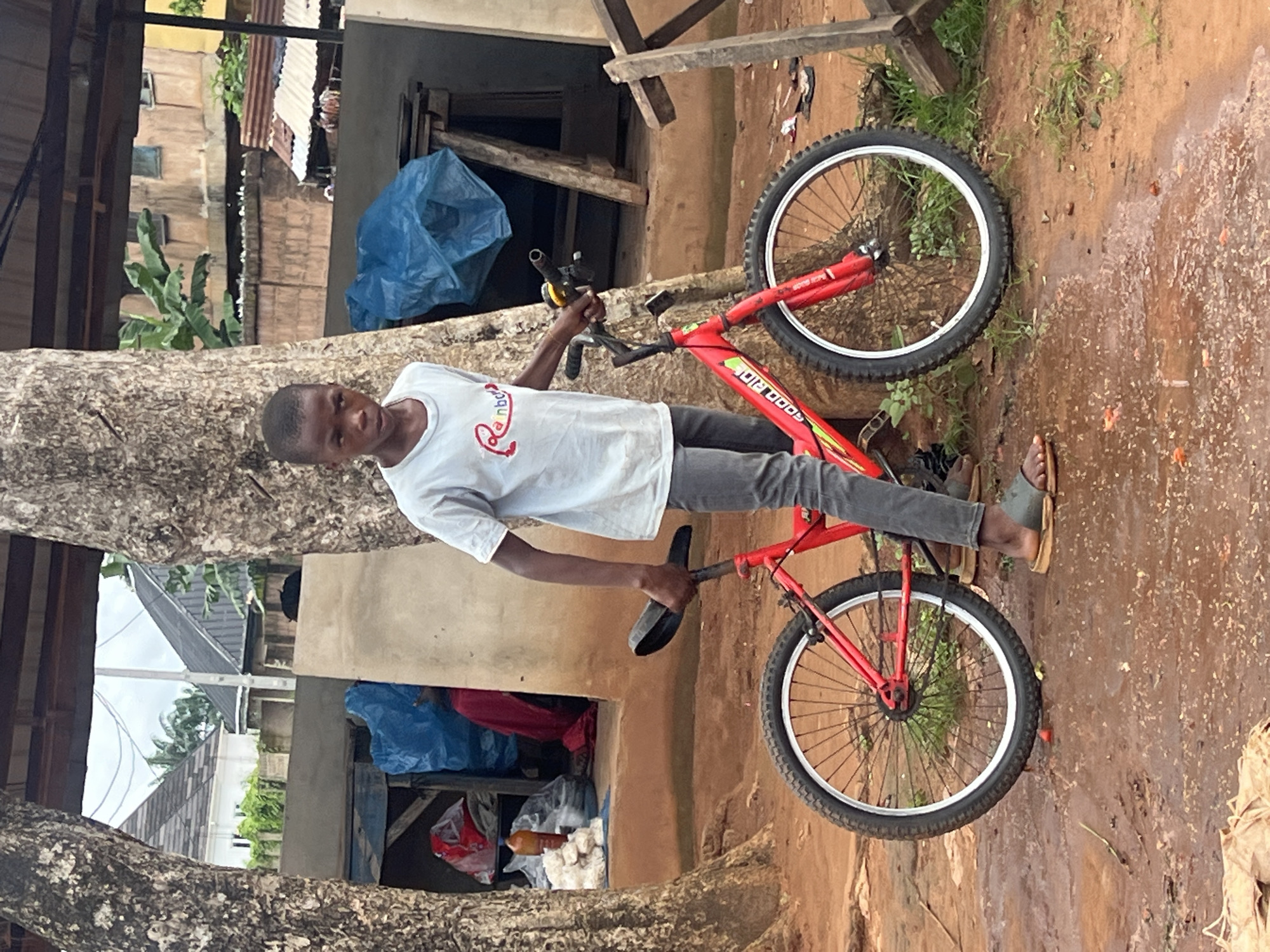 A young Black male stands next to a red bicycle in front of a tree in a yard in Nkpor, Nigeria. The boy is wearing a white t-shirt and grey pants and sandals. The bicycle is red with black tires and silver spokes. The ground is dirt and puddles of water. In the background, there are several buildings and a market stall with blue tarps. The sky is overcast.