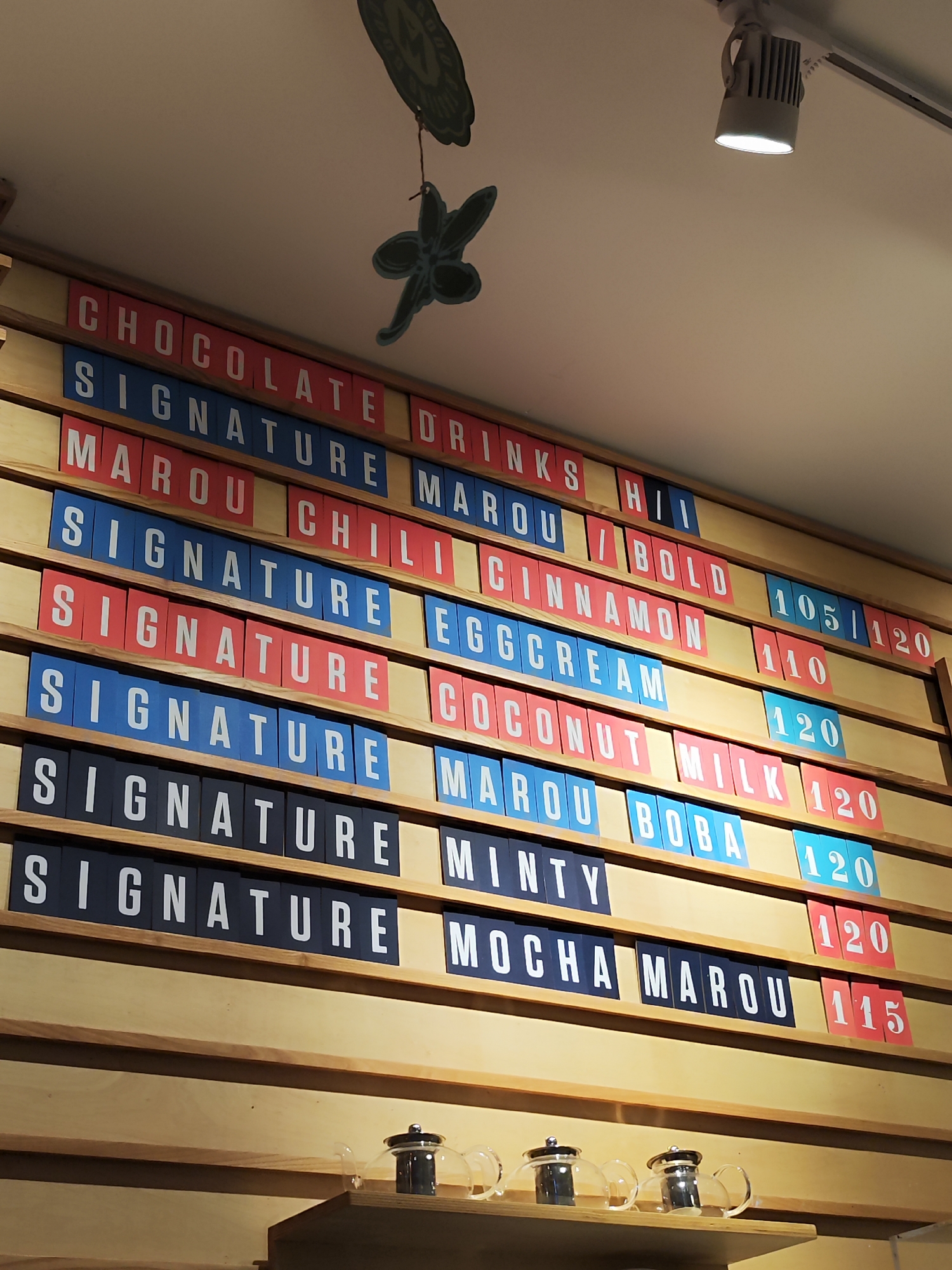 The image shows a menu board on a wooden wall in what appears to be a cafe or shop. The menu is comprised of individual letter tiles in red, blue, and black, arranged in rows to form words and prices. The items listed include "CHOCOLATE DRINKS", "SIGNATURE", "MAROU", "CHILI", "BOLD", "CINNAMON", "EGG CREAM", "COCONUT", "MILK", "BOBA", "MINTY", and "MOCHA MAROU". Prices are shown as numbers in red or blue tiles, such as "105/120", "110", "120", and "115". Two glass teapots are visible on a shelf below the menu. The overall lighting suggests an indoor setting, possibly with overhead track lighting, one of which is clearly visible. There is a decorative item hanging from the ceiling above the menu. The context of the menu being in Nha Trang, Vietnam is not discernible from the image itself.