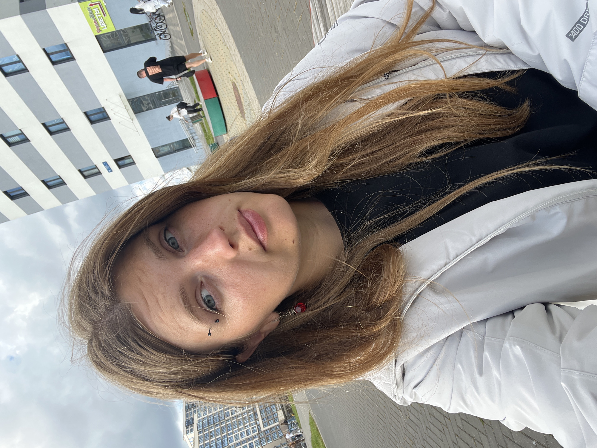 A young woman with long blonde hair and blue eyes is taking a selfie outdoors. She has a black eyebrow piercing and a red earring in her left ear. She is wearing a white jacket over a black t-shirt. In the background, there is a modern, multi-story building with gray and white stripes. Several people are visible in the distance, including two men walking and another person in white. The scene appears to be a public space with paved ground and a small planter box with red and green sections. The sky is cloudy. The image was likely taken in Minsk, Belarus, given the presence of Cyrillic text on a sign attached to the building.
