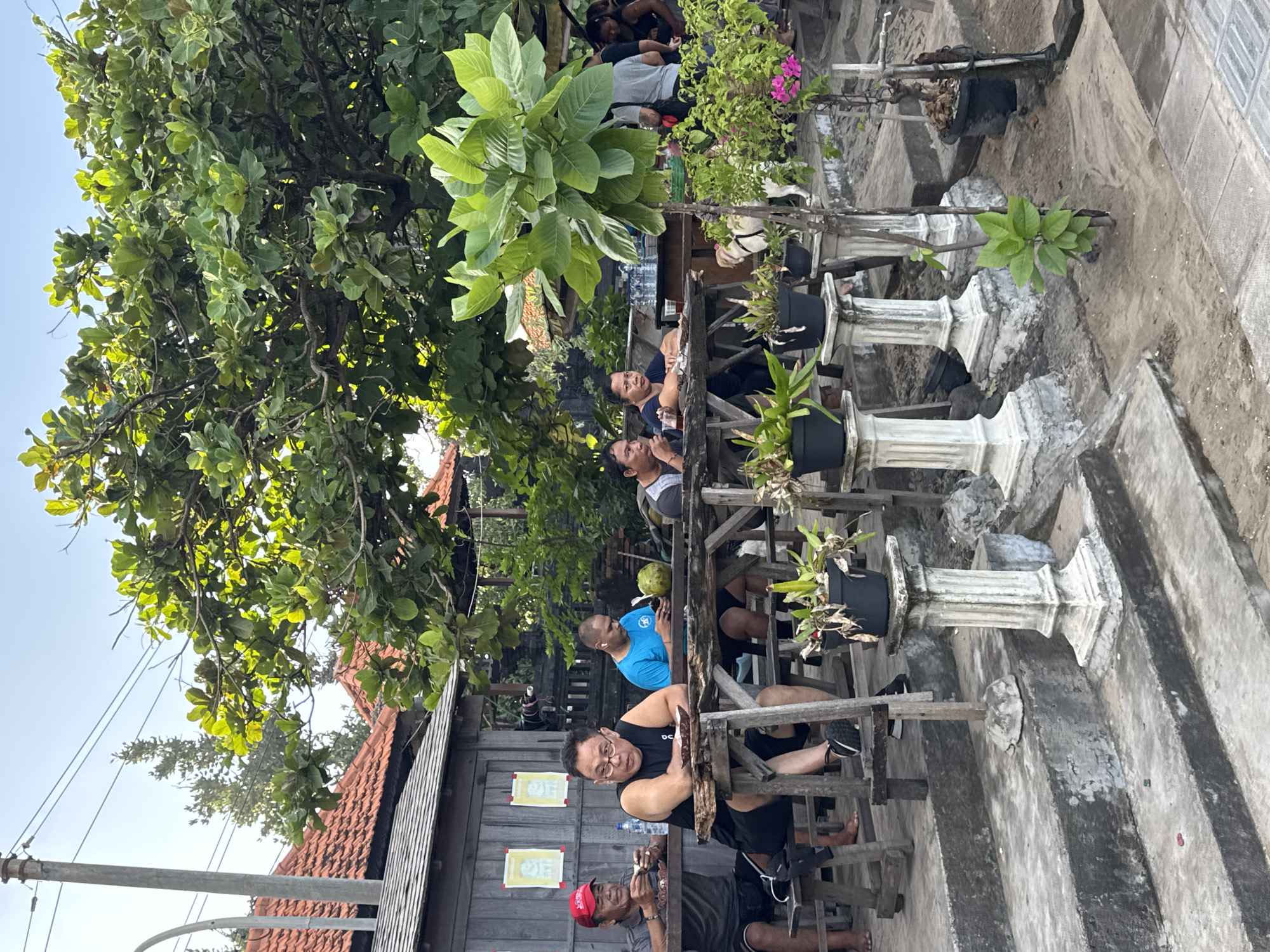 This image captures an outdoor casual dining or gathering area in Kuta, Indonesia. The scene is set under a bright sky with substantial green foliage providing shade.
In the foreground and midground, several individuals are seated at rustic wooden tables with accompanying wooden benches. On the left, a man in a red baseball cap and grey t-shirt is actively eating from a small dish, with a plastic water bottle on the table. Beside him, a man in a black tank top and glasses leans forward, looking towards the viewer, with a small plate on his table. Further right, a man in a blue t-shirt is engrossed with his mobile phone, with a green coconut resting on the table next to him. Another individual is partially visible at a table further to the right. In the deeper midground, two more people are seated at a table, one of whom is looking directly forward. A group of people are standing further back on the right, some appearing to be serving or attending.
The environment features multiple potted plants, some placed on decorative white stone pedestals, enhancing the tropical ambiance. A small white dog, possibly on a leash, is visible between the tables and plants on the right side. The ground consists of concrete steps, sandy patches, and paved areas. In the background, wooden structures with reddish-brown terracotta tile roofs are visible, consistent with local architecture. A prominent utility pole with overhead power lines is situated on the left, extending into the sky. Large, leafy green trees dominate the upper half of the image, contributing to the shaded, outdoor setting. The overall impression is one of a relaxed, informal communal space.