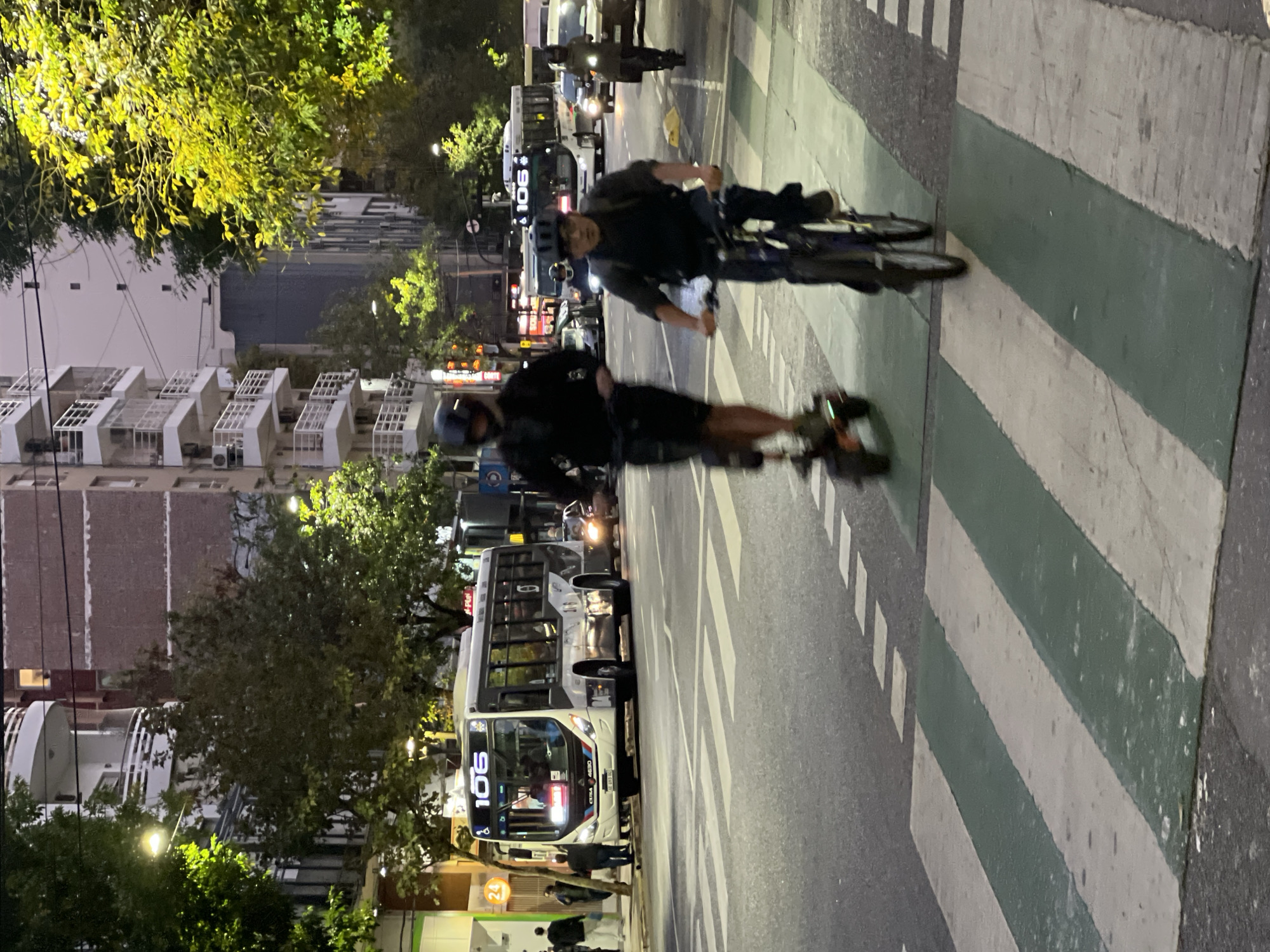 At night in Buenos Aires, Argentina, a person on an electric skateboard and a cyclist cross a crosswalk on a city street in the same direction. They both wear helmets. A 106 bus waits in the street near the curb on the left side of the frame. Apartment buildings and trees line the street behind the bus.