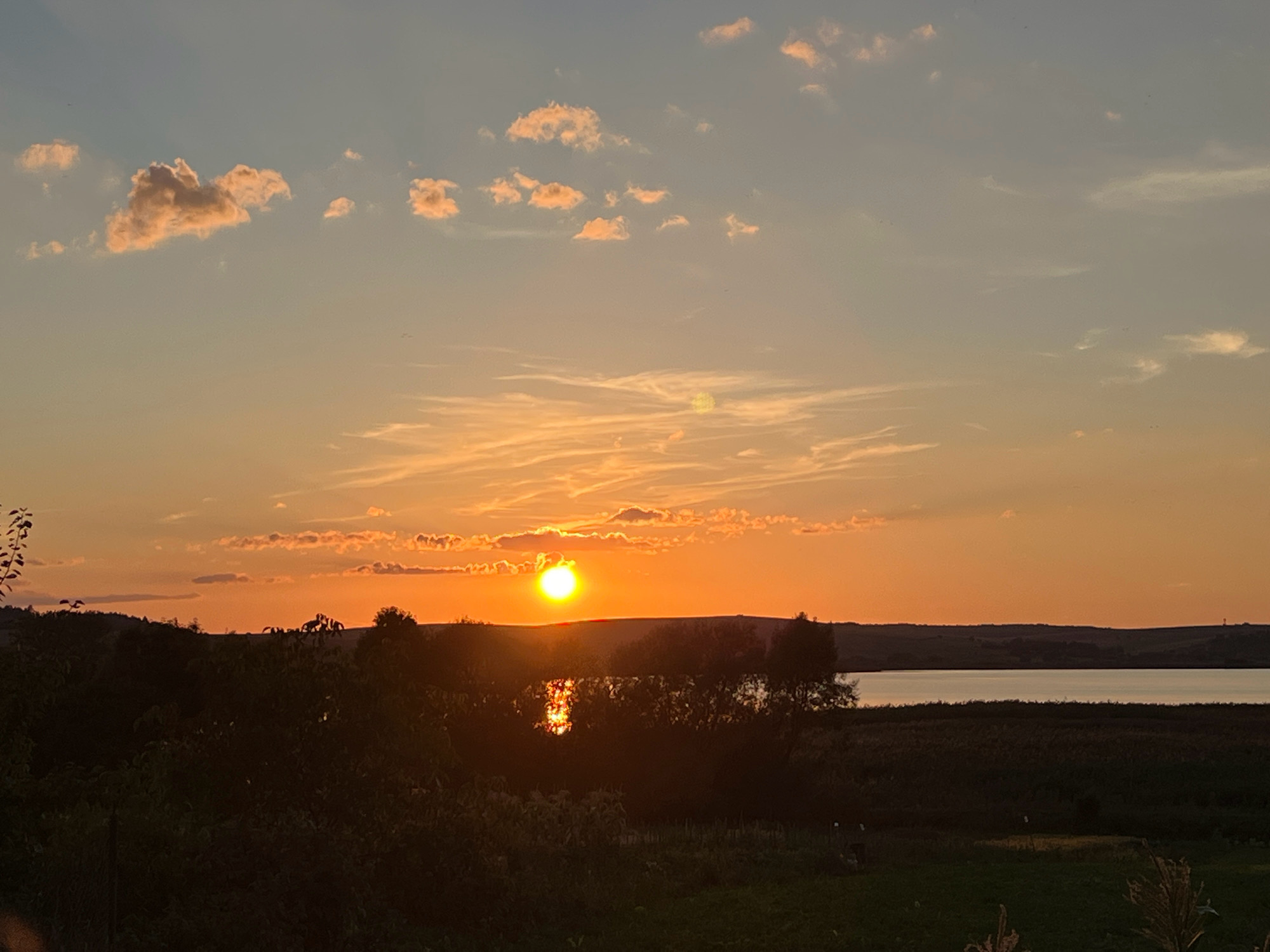 The image captures a serene sunset over a body of water, likely a lake, in the village of Vertelka, Ukraine. The sky is a gradient of soft blues and oranges, with scattered clouds tinged with pink. The sun, a bright orb, is just above the horizon, casting a warm glow and reflecting on the water's surface. In the foreground, dark silhouettes of trees and vegetation frame the scene. The landscape appears rural, with rolling hills visible in the distance. The overall atmosphere is peaceful and tranquil, suggesting a natural, undisturbed environment.