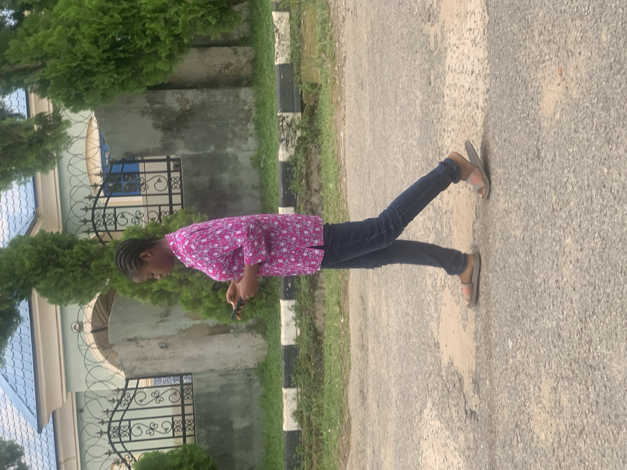 A young Nigerian woman of African descent with dark hair braided is walking to the right while looking down at her mobile phone. She is wearing a pink patterned shirt and dark denim jeans. She has on tan slip-on sandals. She is on an asphalt road, with a grassy area and a black and white striped curb to her left. Behind the curb is a grey wall, a black decorative fence with a curved top, and a building with a blue roof. In the background are green trees. The scene appears to be in a residential area of Abuja, Nigeria, possibly on a street.