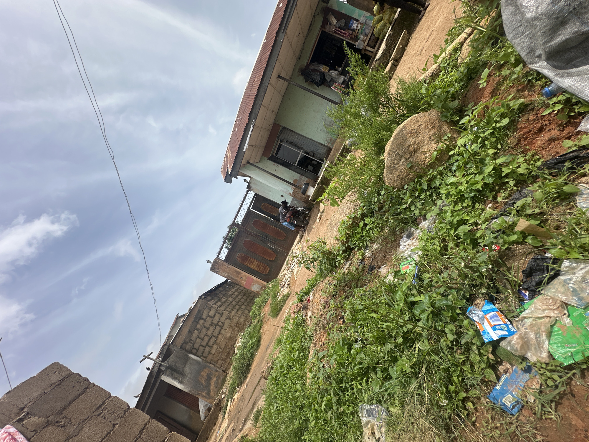 The image depicts a scene in Jos, Nigeria. The setting is an outdoor area with a building that appears to be a shop, characterized by a green painted facade and a corrugated iron roof. In front of the shop, there's a metal gate. A person is visible near the gate, appearing to be on a motorcycle.
The ground is covered in patches of dirt and lush green vegetation, with a large rock in the foreground. There is also a notable amount of scattered litter, including plastic bottles and wrappers, contributing to a sense of disarray in the immediate surroundings. A grey tarp-like object is also visible on the right side of the frame.
In the background, other structures are present, including buildings made of concrete blocks. The sky above is mostly overcast with some visible clouds and wires, suggesting it is daytime. The overall atmosphere conveyed by the scene appears to be that of an everyday, perhaps somewhat neglected, urban or semi-urban environment. There is no discernible text in the image.