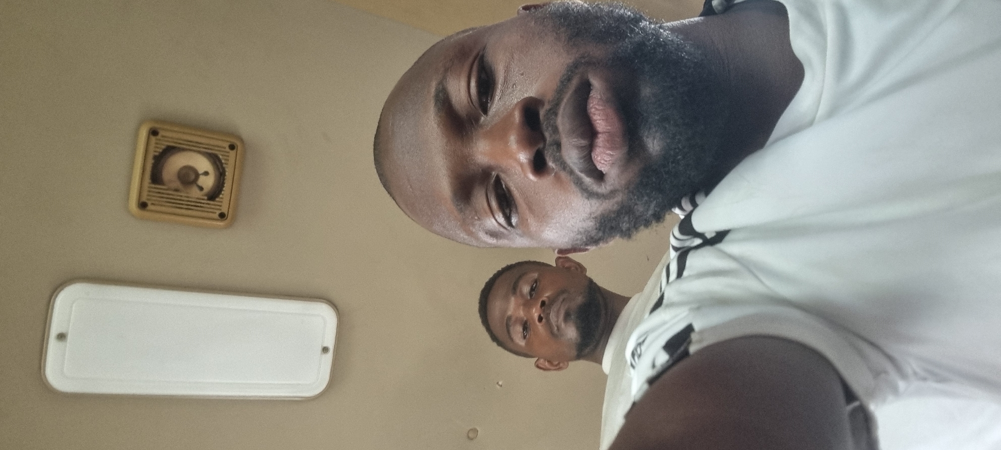 The image, taken in Ilishan, Nigeria, features two men captured from a low-angle perspective, looking down towards the camera. The primary subject in the foreground is a man with dark skin, a full black beard, and short dark hair. He has a neutral expression, with his lips slightly parted, and his eyes are directed at the lens. He is wearing a white shirt, visible on the right side, which appears to have some dark striping or patterns on the sleeve area.
Behind him, positioned towards the lower right, a second man is partially visible. He also has dark skin and a beard, and his gaze is directed downwards at the camera with a neutral expression. He is wearing a white shirt.
The background consists of a plain, light-colored surface, likely a ceiling or an upper wall. Mounted on this surface are two distinct objects. In the upper left corner, there is a square, light-beige fixture with a circular, gridded opening in its center, containing a small, three-pronged element. This object resembles a small speaker or a ventilation unit. Below and to the left of this, a longer, rectangular white fixture is visible, consistent with a light cover or another type of ceiling vent.
The photograph appears to be an indoor shot, possibly a selfie or a photo taken from a very low vantage point. No visible text is present in the image, and the time of day is not discernible.