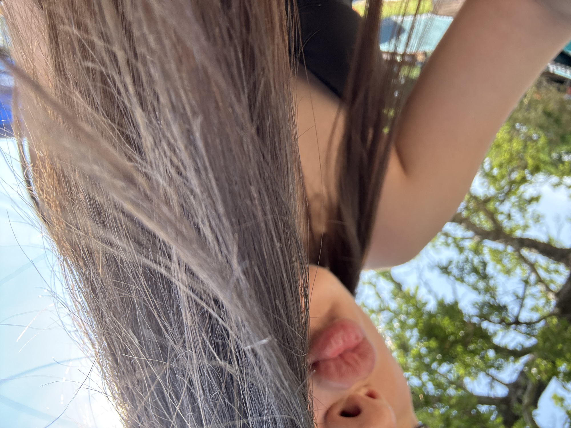 A low-angle, close-up photograph captures the upper body of a person, likely female, partially obscured by their own hair. The subject displays light skin tone and long, straight brown hair that sweeps across the foreground, appearing illuminated by sunlight. Visible facial features include the lower portion of the nose and prominent, pursed lips, formed into a "kissing" expression. A segment of the left shoulder and upper arm is visible, clad in a dark, sleeveless garment. The background is an out-of-focus outdoor environment, featuring blurred green foliage from trees on the left and a bright, clear blue sky in the upper right. Natural sunlight illuminates the scene. The specific location in the city of Kostievo, Bulgaria, cannot be determined from the visual information present in the image.