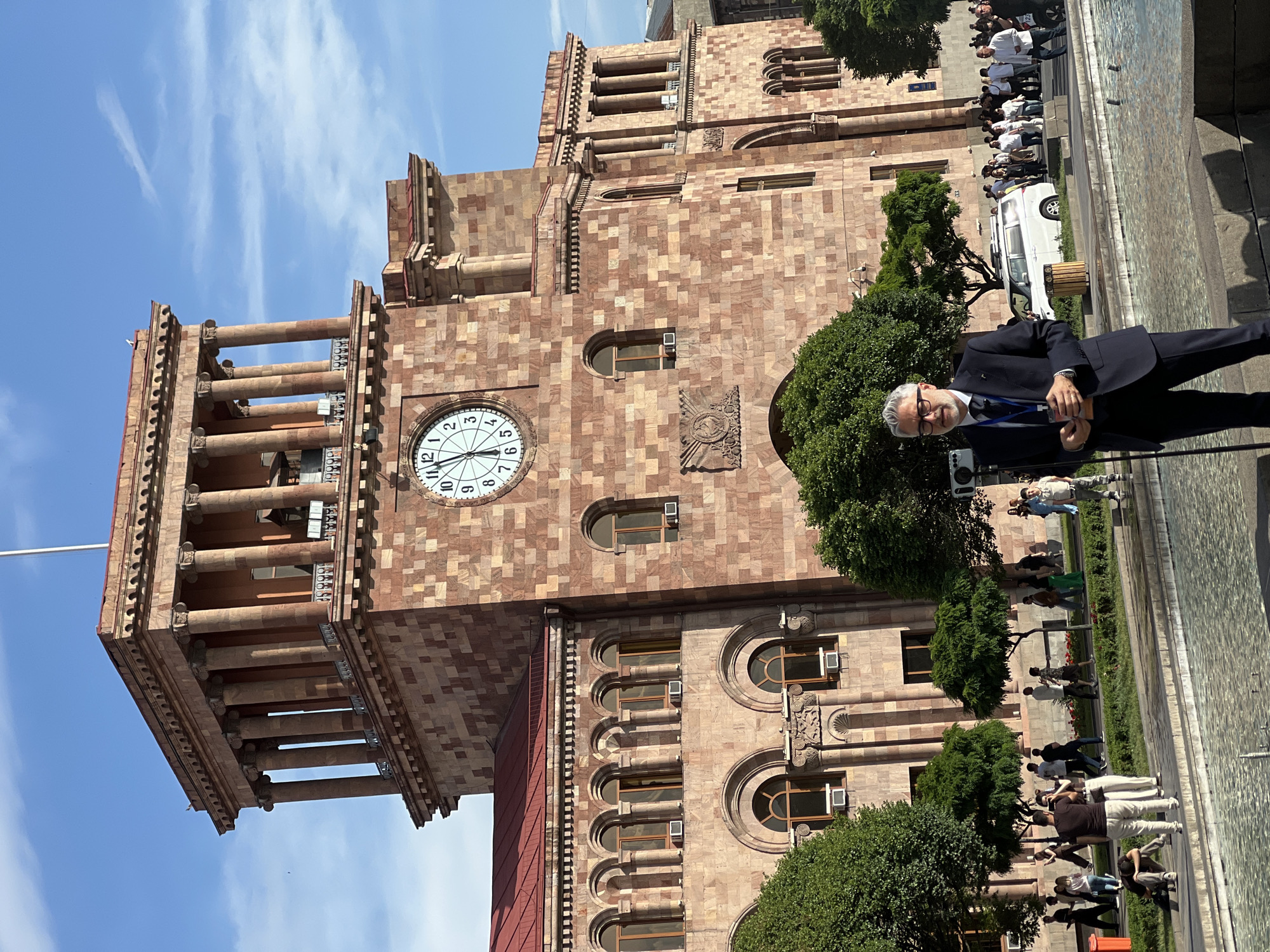 This image captures a vibrant public scene in Yerevan, Armenia, dominated by a magnificent, multi-story civic building. Constructed from striking reddish-brown and lighter stone blocks, likely local tuff, the architecture features a distinctive mosaic-like pattern. The building boasts numerous arched windows, elaborate decorative carvings, and a prominent circular clock on its facade. The clock, displaying Roman numerals, indicates a time around 11:30 or 11:35. To the left, a grand clock tower rises, characterized by ornate columns and balustrades.
In the immediate foreground, a distinguished man with gray hair and a beard, wearing glasses, a dark suit, light blue shirt, and tie, stands looking towards the viewer. He holds what appears to be an electronic device or ID badge attached to a blue lanyard. His demeanor is composed and neutral.
Behind him, a bustling public area unfolds, with a wide paved space, possibly a square or promenade. Numerous people are present, some casually walking, others gathered in groups. A noticeable line of individuals, many dressed in white shirts, suggests an organized event or assembly. A white van is partially visible amidst the crowd in the middle distance. Lush green trees and shrubs line the front of the building and the paved area, contributing to the urban landscape. A dark, reflective surface on the right could be a fountain or reflecting pool.
The scene is bathed in bright, clear sunlight under a blue sky adorned with faint wispy clouds, indicating a sunny day, likely during midday or early afternoon. The impressive architecture, combined with the presence of a crowd and a man in formal attire, suggests a significant public occasion or daily activity in a prominent city location. No legible text beyond the Roman numerals on the clock face is discernable.