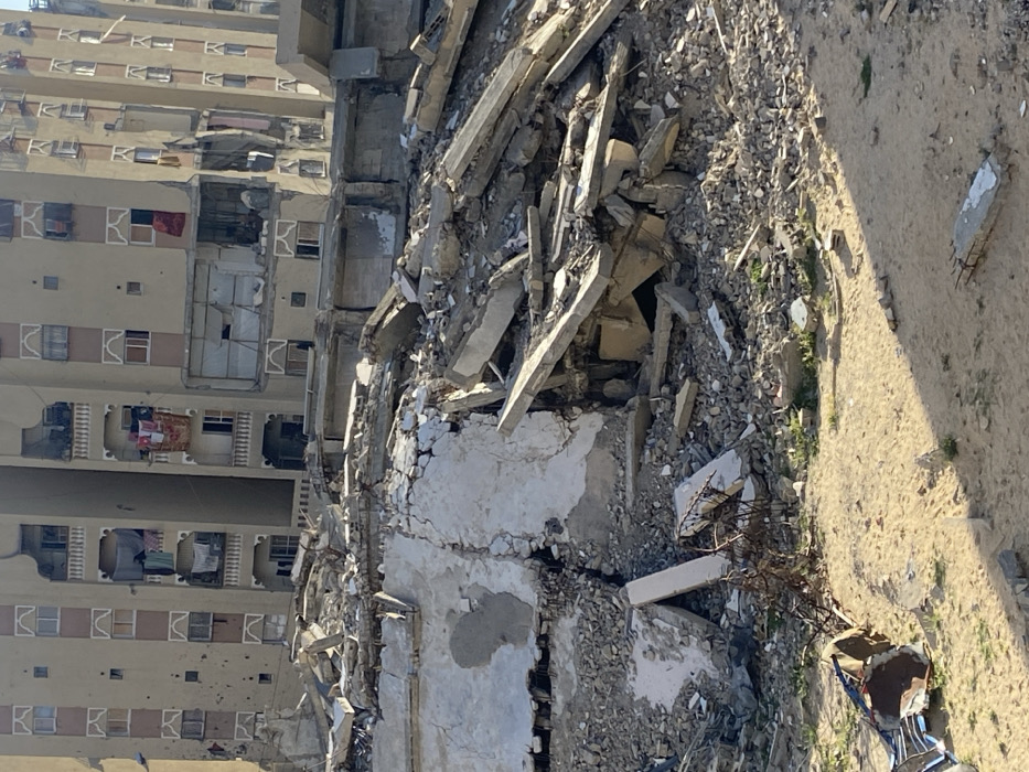 The image depicts a scene of extensive destruction in Khan Yunis, Palestinian Territory, under clear daylight. The foreground and midground are dominated by a massive, irregular mound of rubble, consisting of shattered concrete slabs, exposed rebar, broken masonry, and pulverized building materials. Large, fragmented concrete sections are visibly tilted and stacked. The immediate ground level is sandy soil with sparse patches of green vegetation. To the lower left, a twisted metal structure lies on the sand, alongside other debris like corrugated metal. In the background, two multi-story residential buildings are visible. The closer building, of light-colored construction, exhibits significant damage, including large structural breaches and shattered windows on its lower and middle floors. Numerous small impact holes are visible across its facade, and laundry or fabrics hang from several balconies. The adjacent building further to the right also shows signs of damage. No people are present in the visible areas of the scene.