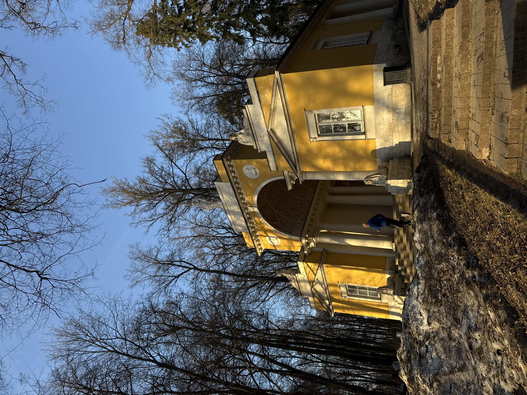 Here's a detailed description of the image:
**Scene:**
* A bright yellow, classical-style building is prominently featured, with white columns and arched doorways. The architecture suggests a historical structure, possibly a pavilion or small palace. The building is set against a backdrop of bare trees, indicating a fall or winter season. A blue sky is visible through the branches.
**Objects:**
* Stone steps lead up to the building's entrance. Some snow or ice is visible on the ground near the steps.
**People:**
* A man is standing on the steps of the building. He is wearing a blue jacket.
**Environment:**
* The lighting suggests it is either early morning or late afternoon, with long shadows cast across the scene. The setting appears to be a park or garden due to the presence of trees and open space.
**Location:**
* The image is of the Pavilion "Temple of Ceres" in Tsaritsyno Park in Moscow, Russia.