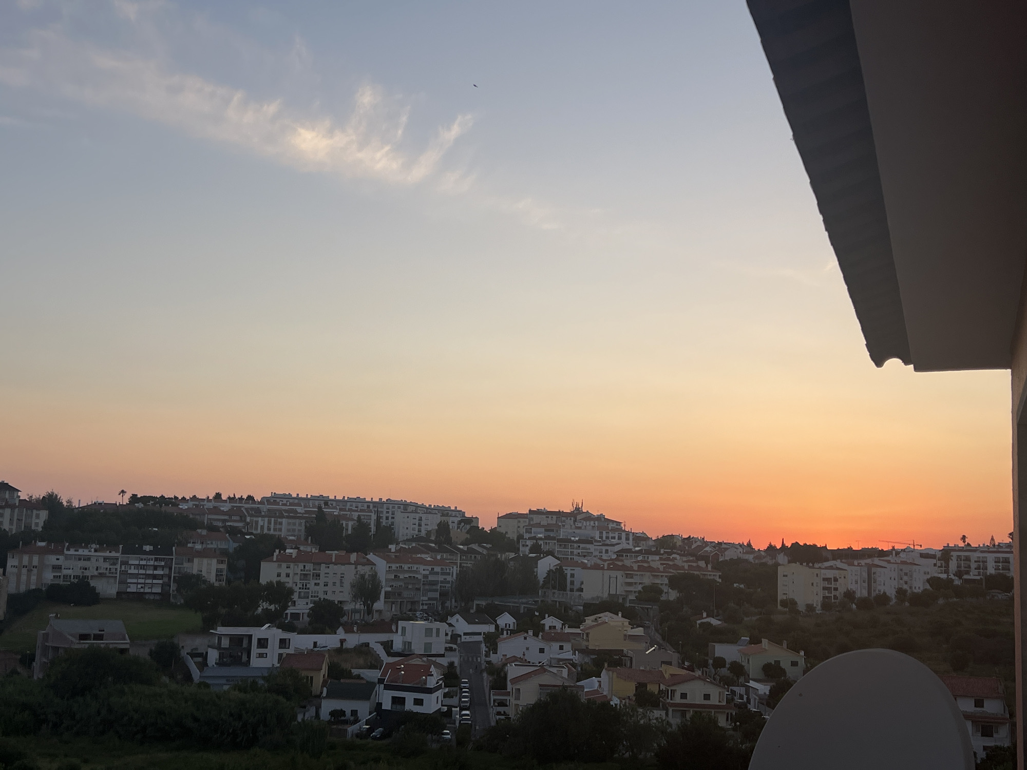 An aerial wide shot captures a residential area of Cascais, Portugal, during a sunset. The sky transitions from a pale blue at the top to a vibrant orange and pink hue near the horizon. A sliver of the roof of a building juts in from the top right corner of the frame. The cityscape features a mix of apartment buildings and houses with red-tiled roofs, interspersed with lush green trees and vegetation. A faint wispy cloud formation is visible in the upper left quadrant of the sky, and a distant bird is a small black speck in the upper middle. A satellite dish occupies the lower right portion of the image, partially obscuring the view of the houses below. The overall impression is one of a peaceful, urban evening.