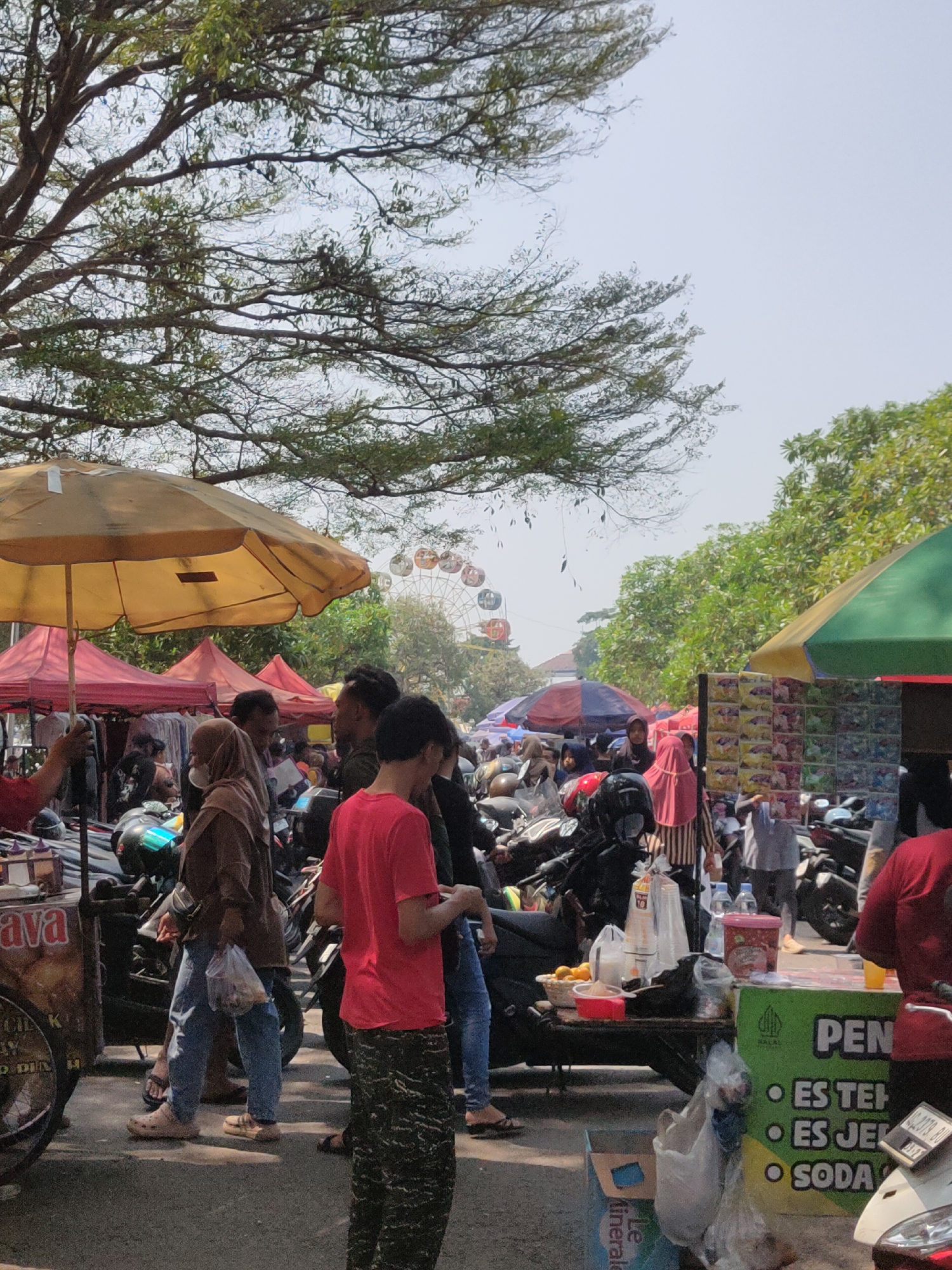 This is a vertical, outdoor, medium-shot photograph taken in the city of Ngasem, Indonesia, capturing a bustling market scene. The foreground features a food stall with a green and yellow facade displaying text that reads "PEN ES TEH ES JUS ES SODA". Various items are arranged on a table in front of the stall, including plastic cups, bottles of water, oranges in a red bowl, and bags of groceries. A motorcycle is parked to the right, and a blue cardboard box with text is on the ground.
In the mid-ground, people are seen milling about, some wearing face masks. A person in a red t-shirt is standing to the right, facing away from the camera. To the left, a person in a brown jacket and blue jeans is holding a clear plastic bag. Several other individuals, some in headscarves, are visible among stalls and parked motorcycles. Overhead, large tree branches with sparse leaves create a canopy, with a Ferris wheel visible in the background through gaps in the foliage. Red and yellow market stalls with peaked roofs are set up, and a yellow umbrella is prominent in the left portion of the image. The ground is paved with asphalt. The overall atmosphere suggests a lively daytime market.