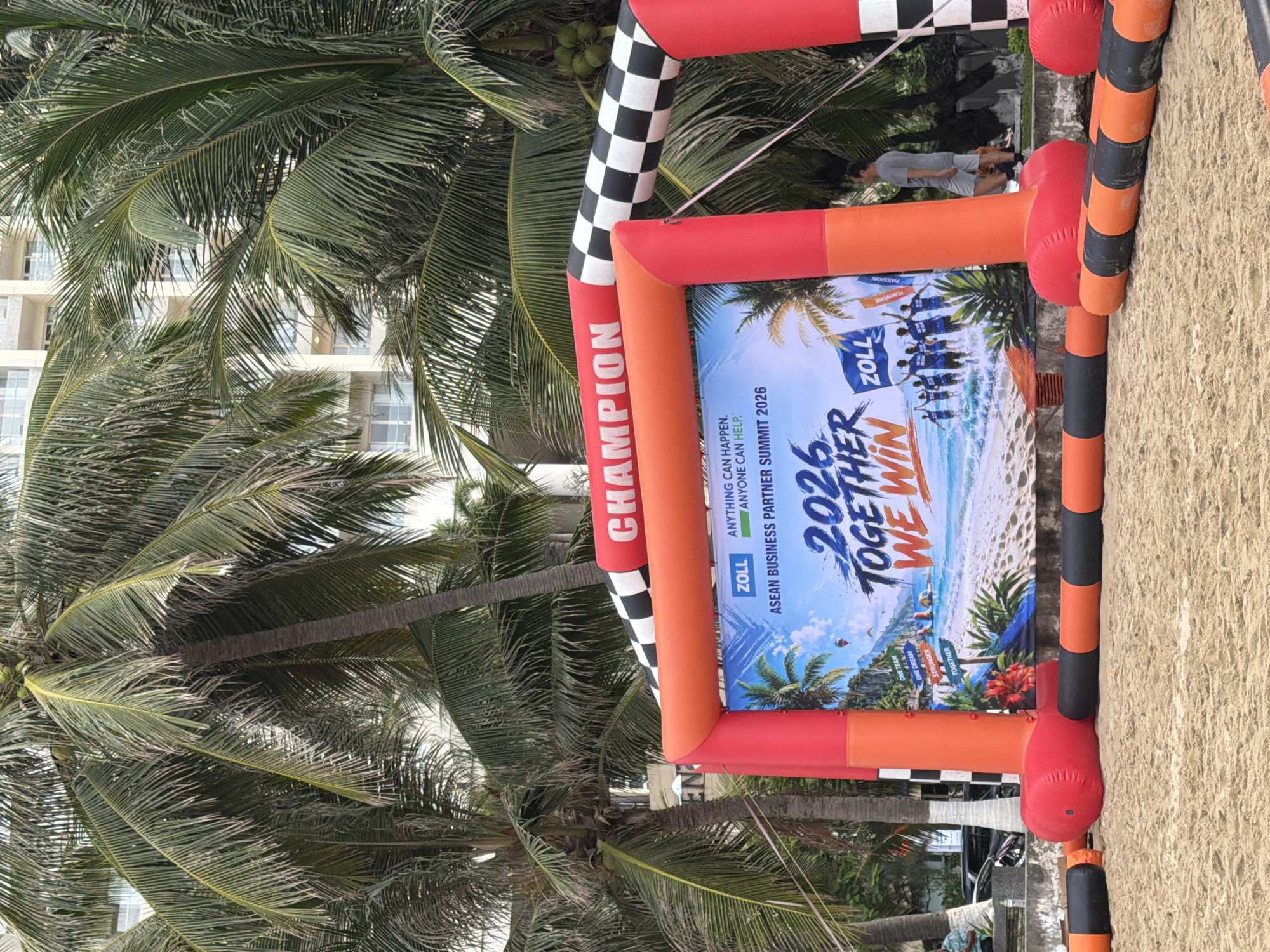 The image depicts an outdoor event setup on a sandy beach, likely in Nha Trang, Vietnam, as suggested by the presence of palm trees and a modern building in the background. In the foreground, an inflatable archway with red and orange elements stands prominently, featuring a checkered flag pattern at the top and the word "CHAMPION" in white text. Below the arch is a large digital screen displaying a beach scene with the text "ASEAN BUSINESS PARTNER SUMMIT 2026" and "2026 TOGETHER WE WIN." Several inflatable barriers, alternating in black and orange, are laid out on the sand, forming a path or boundary.
Behind the archway, lush green palm trees dominate the scene, their fronds reaching towards a pale sky. In the middle ground, a light-colored, multi-story building with numerous windows is visible, partially obscured by the palm trees, suggesting a hotel or resort common in coastal cities. A few people are present: one man in a gray t-shirt and shorts stands near the inflatable arch, looking towards the screen. Another person in a blue shirt is partially visible to the left, sitting down. In the distance, a black car is parked, and other individuals are seen walking or standing further back, beyond the event setup. The overall environment is that of a sunny beach event, possibly a race start or finish line, with clear sand and tropical foliage.