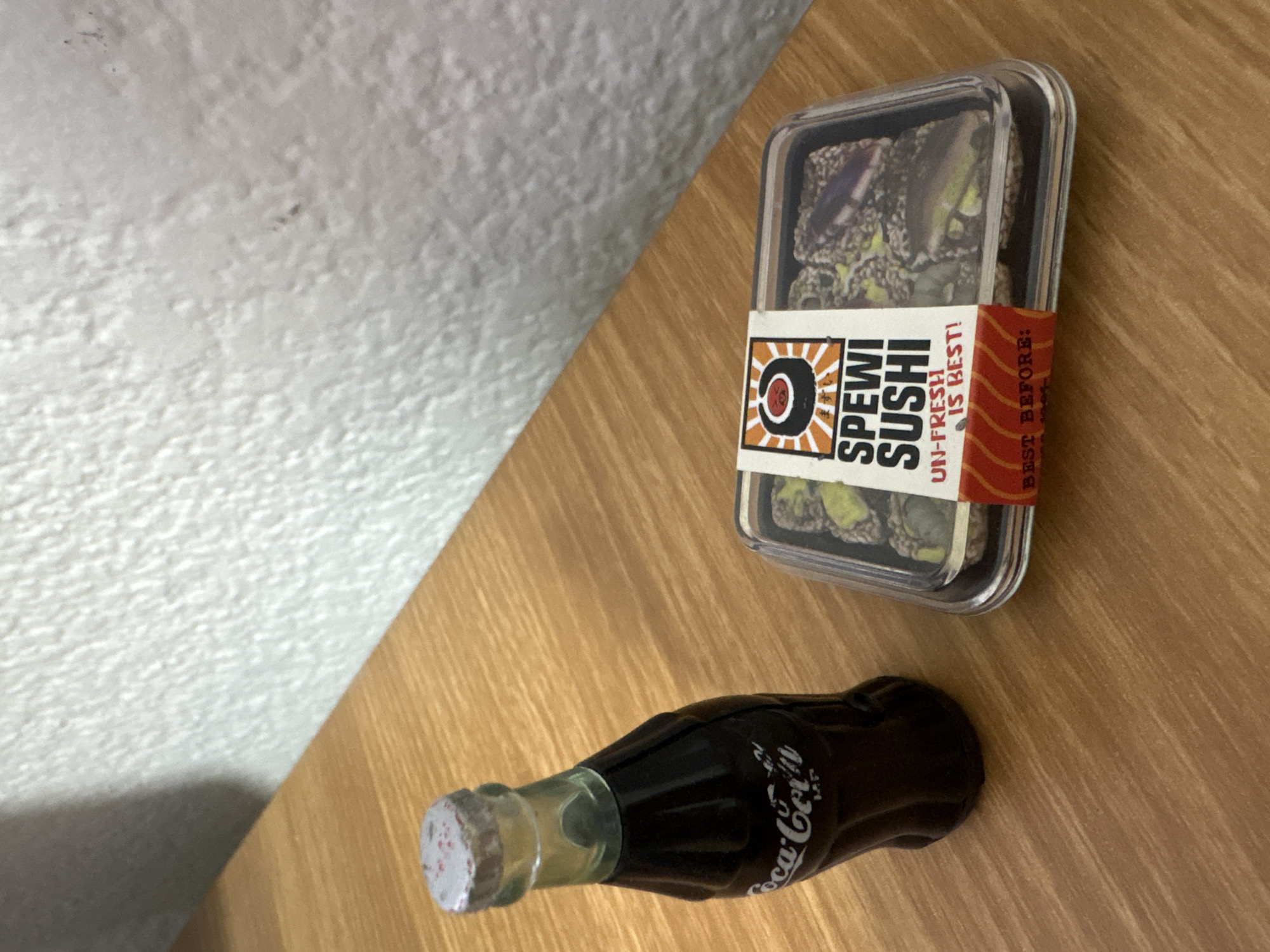 A plastic container of "Spewi Sushi" with a label that reads "UN-FRESH IS BEST!" sits next to a miniature Coca-Cola bottle on a wooden surface. The scene has a white textured wall in the background. There is no evidence to determine the location is in Cuernavaca, Mexico.