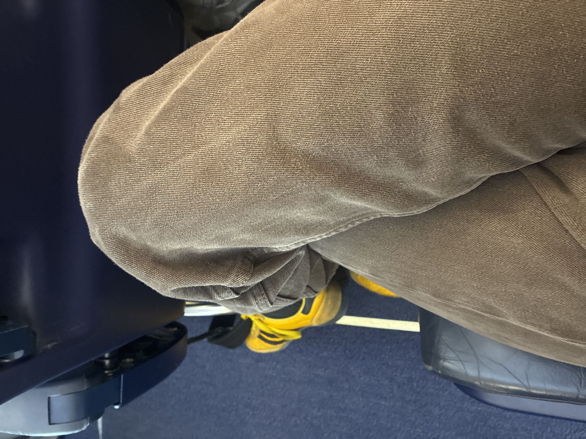 A close-up, high-angle shot depicts a seated person with knees bent. The individual is wearing brown corduroy pants and yellow sneakers with black stripes. The setting appears to be an airplane cabin, suggested by the blue seat and armrest on the left. A seat is also visible to the right. The location, Sant Josep de sa Talaia, Spain, cannot be definitively determined from the image alone, but is stated as given context.