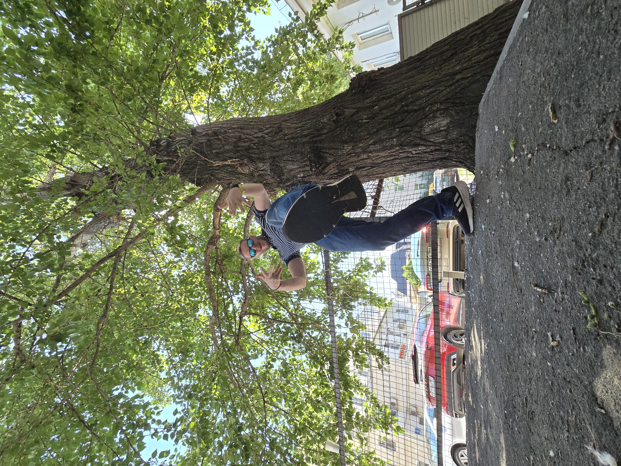 A man, wearing sunglasses, blue jeans, and a striped shirt, is standing next to a tree trunk, holding a black skateboard. The tree is in front of a wire fence, and there are buildings and parked cars visible in the background. The scene is in Moscow, Russia. The photograph appears to be taken from a low angle, rotated 90 degrees clockwise.