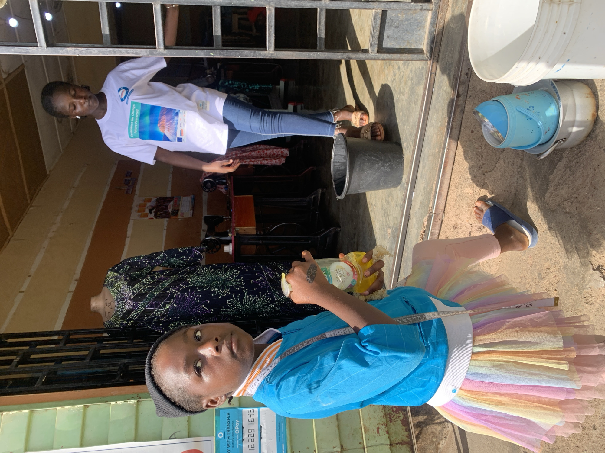 A medium shot depicts two young individuals in an urban environment, possibly within the city of Jos, Nigeria. The foreground features a child wearing a black beanie, a blue shirt, and a multi-colored pastel tutu skirt. A measuring tape is draped around their neck. They hold a bottle labeled "Organic Honey Hand Wash." In the background, a woman is seen in a doorway, wearing a white t-shirt with a blue design. A sewing machine and a mannequin are visible inside the building. There are several plastic buckets to the right of the frame. The scene is bathed in sunlight. A sign with the OPay logo is located near the child.