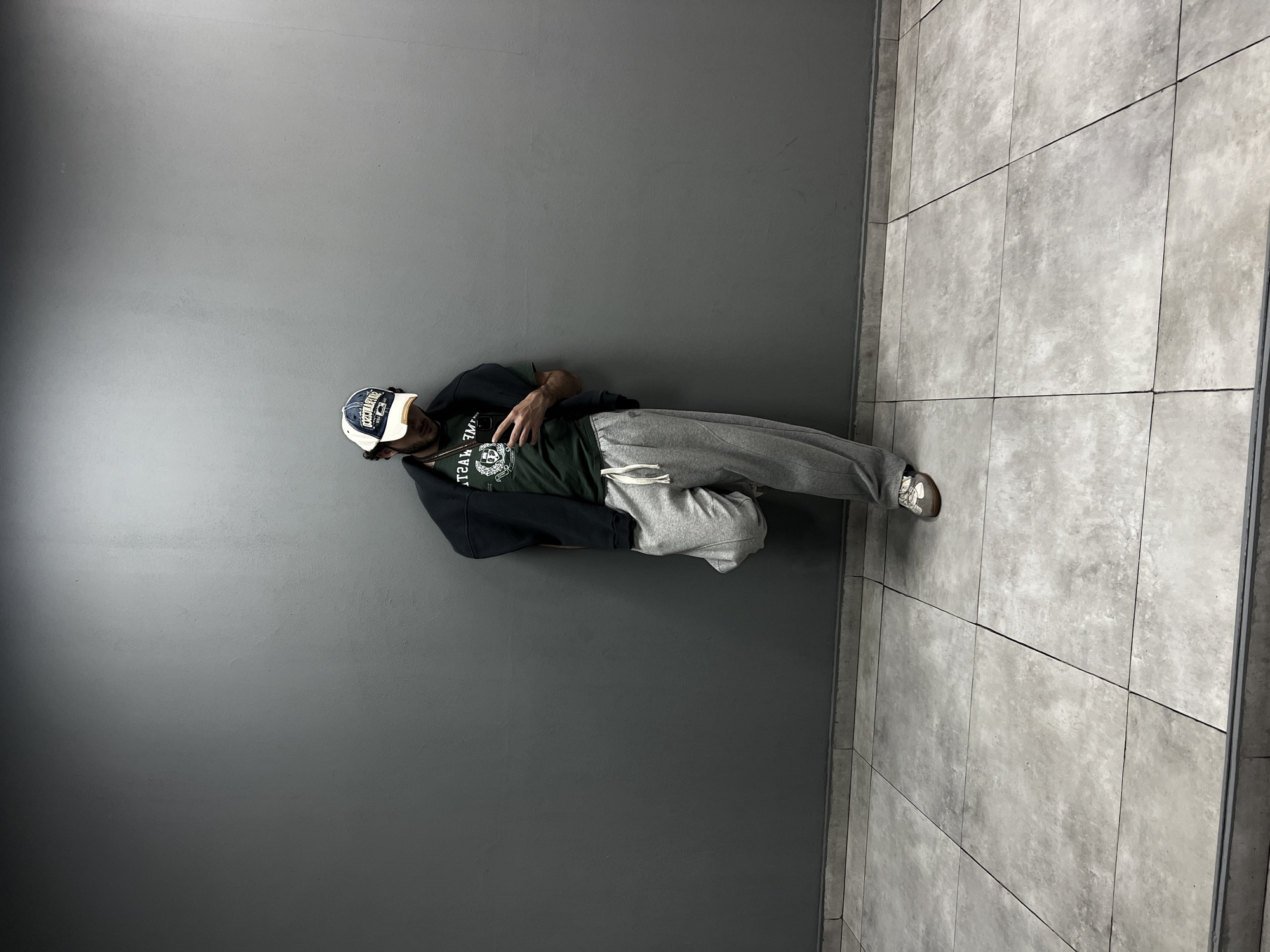 A man is leaning against a gray wall, with the lower half of his body extending out onto a tiled floor. He is wearing a baseball cap, a black hoodie over a green t-shirt, and gray sweatpants with sneakers. The environment consists of a plain, gray wall adjacent to a light gray tiled floor with dark grout lines. The specific location in Yerevan, Armenia cannot be determined from the visual information alone.