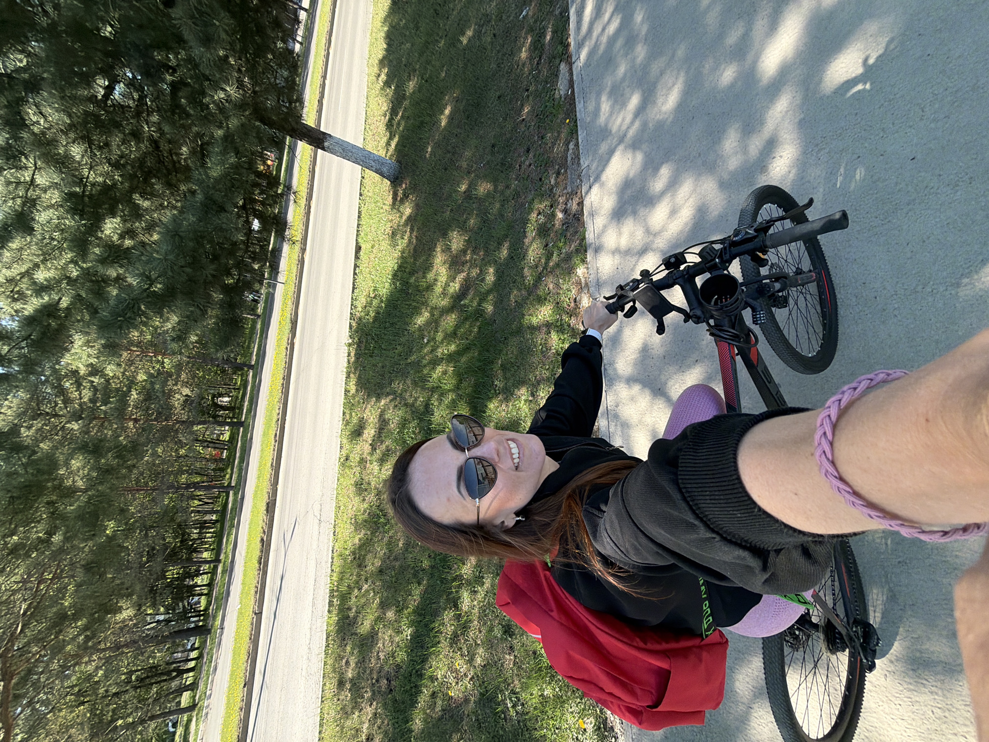 A woman is taking a selfie while riding a bicycle along a paved path. The path is bordered by a strip of grass and a road with trees lining the other side. The woman is wearing sunglasses, a dark-colored jacket, and leggings, and has a red bag on her back. She is smiling at the camera. The location appears to be a park or green area within a city. Although the prompt mentions Birobidzhan, Russia, there is no information in the picture to confirm or deny that it is the location.