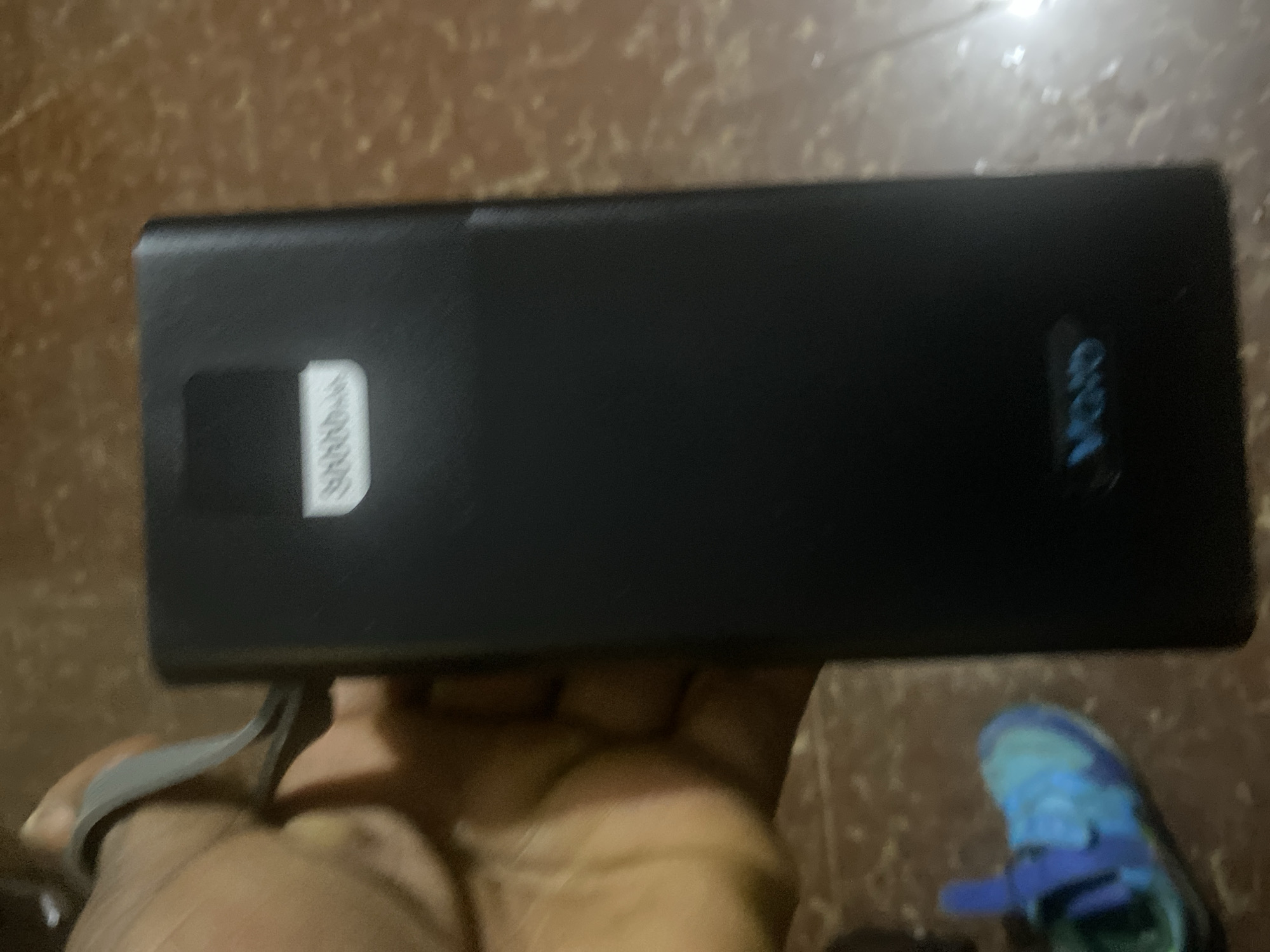 In the city of Jos, Nigeria, a light-skinned hand holds a black, rectangular electronic device. The device displays "20000mAh" on a white label near its top and a blue glowing "VORO" logo at its base. A grey strap or band is visible around the wrist of the hand. In the lower-left foreground, a blue and green athletic shoe rests on a textured, brownish-orange floor. The ambient lighting is dim.