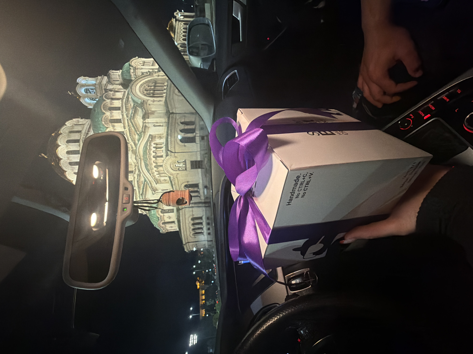 The image, taken from inside a car at night, shows a white, square gift box with a large purple ribbon on the passenger seat. The box has text on its side that reads "Handmade. No CTRL+C, No CTRL+V." A person's hands are visible near the box and on the car's dashboard. The car's rearview mirror is visible in the frame.
The background visible through the windshield shows the Alexander Nevsky Cathedral, a major landmark in Sofia, Bulgaria. Streetlights and other buildings are visible in the city beyond the cathedral.