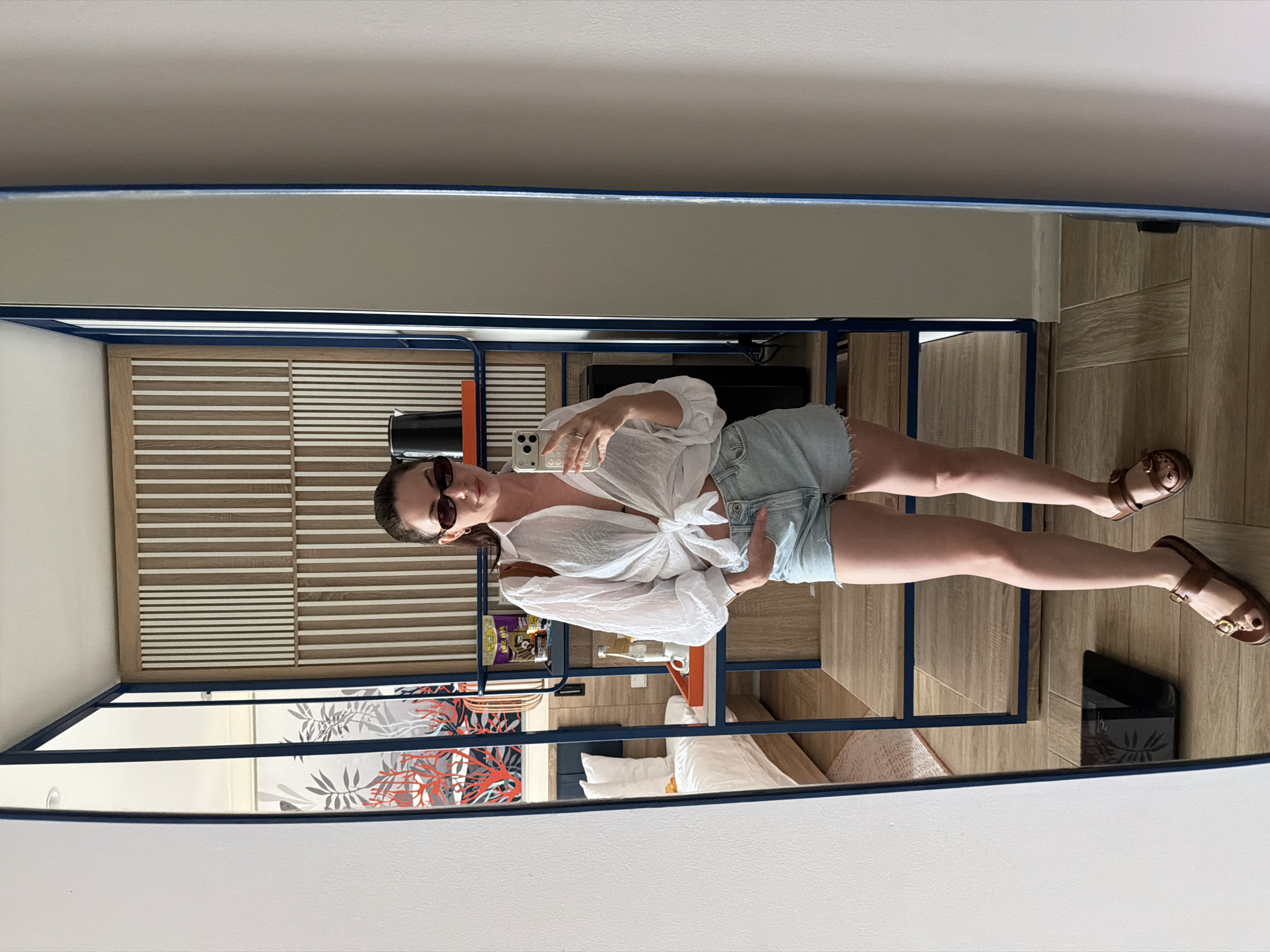 A woman is depicted taking a mirror selfie within a room in Dong Ninh Hoa Ward, Vietnam. The woman, appearing in her 20s or 30s, wears dark oval sunglasses, a sheer white long-sleeved shirt tied at the waist over a dark top, light-wash denim shorts with frayed hems, and brown sandals with gold buckles. She holds a white smartphone with multiple rear cameras in her right hand, with her left hand in her shorts pocket. A ring is visible on her right hand's ring finger, and her toenails are painted dark.
The mirror, framed in dark blue, reflects a room featuring a bed with white bedding and a blue headboard to the left. A bedside table with an orange tray and a patterned rug are adjacent to the bed. The wall above the bed displays a mural with red coral and grey plant motifs on a white background. To the right of the woman's reflection, a multi-tiered wooden shelving unit with a dark blue frame is visible. The top shelf contains a black kettle and an orange-spined object. The middle shelf holds packaged items, a water bottle, and a white cup. A slatted wooden and white wall panel is behind the shelving unit. The floor is covered in light-colored tiles. A black digital weighing scale is on the floor to the left of the woman's feet in the foreground, reflected in the mirror. The walls surrounding the mirror are plain and light-colored.