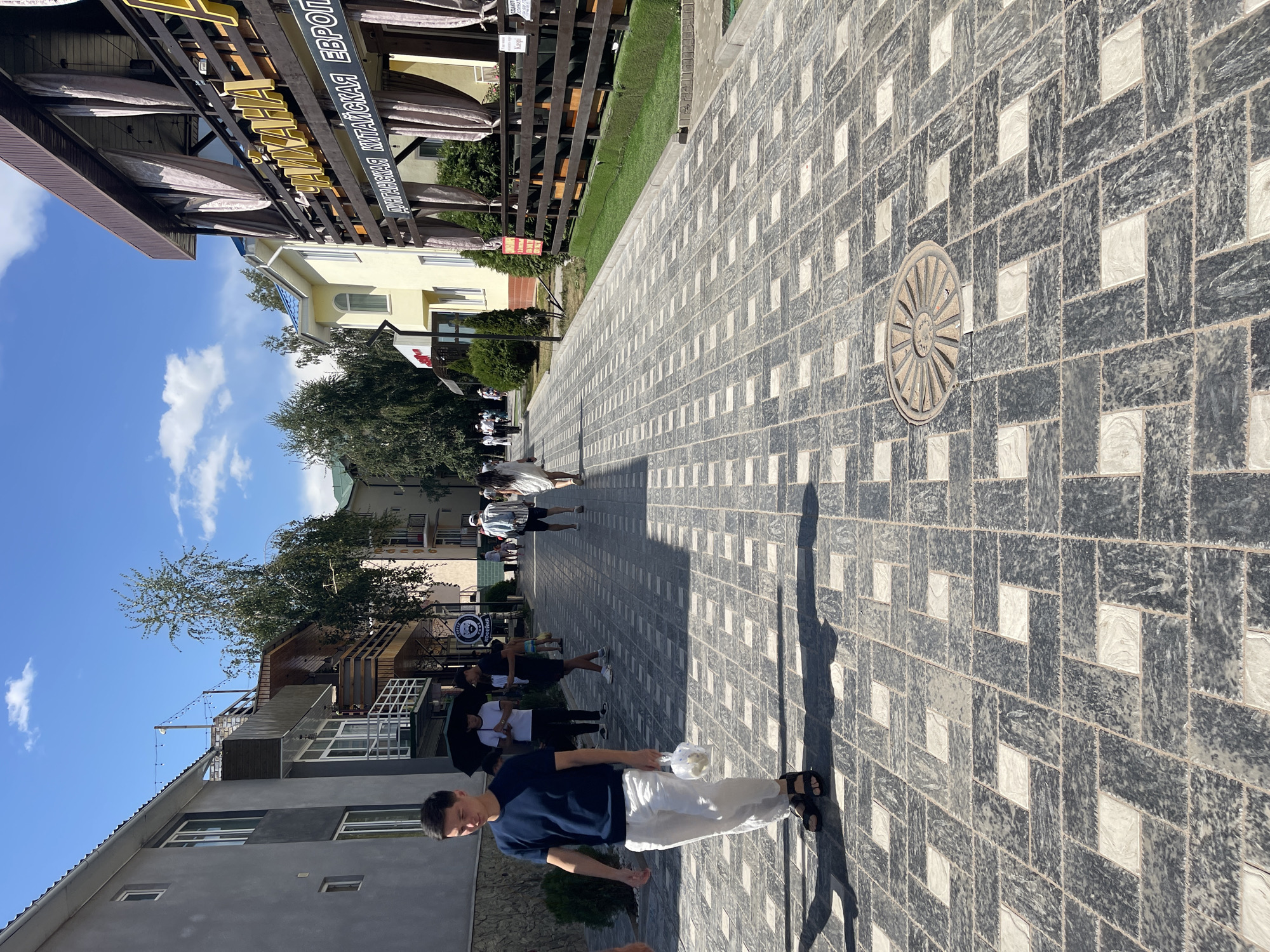 A street scene in Bosteri, Kyrgyzstan, shows a pedestrian walkway paved with a patterned cobblestone surface. On the left, a man in a dark blue t-shirt and white pants walks towards the viewer, carrying a white bag in his right hand. He is wearing black sandals. Behind him, several other individuals are visible, some with umbrellas. Buildings line both sides of the street. The building on the right features a second-story balcony with curtains and signage that reads "КАЙХАНА" and "ДУНГАИСКАЯ КИТАЙСКАЯ ЕВРОП". The sky above is blue with scattered clouds. Trees are present along the walkway and behind the buildings. A circular manhole cover is embedded in the cobblestones in the mid-ground. The overall impression is of a sunny day in a populated area, likely a tourist or commercial street.