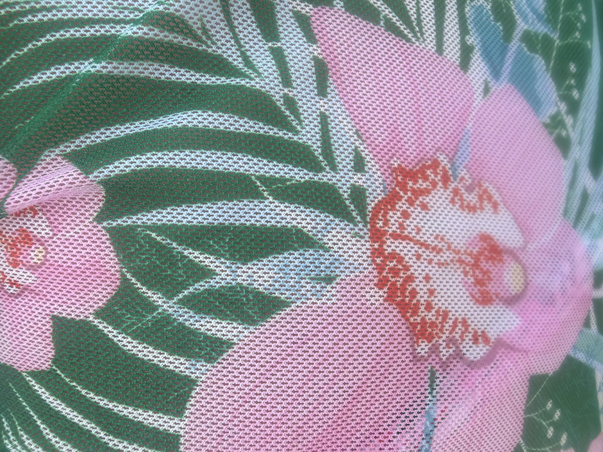 This is a close-up, abstract shot of a patterned fabric. The fabric has a mesh-like texture, creating a pixelated effect on the design. The pattern features large, stylized floral elements in shades of pink and white, with accents of red. Interspersed with the flowers are broad, green leaf shapes, outlined by thin white lines. Hints of light blue and darker green are also visible, contributing to a tropical or botanical theme. The lighting is diffused, suggesting it might be an overcast day or an indoor setting. No people, specific objects, or environmental cues indicating a location are discernible beyond the fabric itself. There is no visible text. The overall impression is one of vibrant, decorative textile art.
