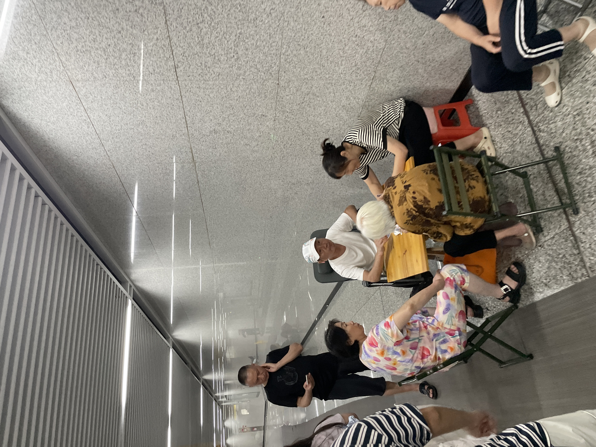In an indoor walkway, possibly a subway station or transit hub in Liangjiang, China, several people are gathered. A man in a black t-shirt and dark shorts is standing and holding a mobile phone, looking downwards. In the foreground, a woman wearing a colorful floral-patterned outfit is seated on a green folding chair. Facing her, a man in a white t-shirt and a white baseball cap is seated at a small wooden table. Another woman, dressed in a black and white striped shirt, is also seated at the table, leaning forward. To the right, partially visible, a person in dark clothing with white stripes on the pant legs is seated. The environment features a textured grey wall and a slatted ceiling with fluorescent lighting. The floor is a mottled grey tile. Some of the individuals appear to be engaged in conversation or playing a game at the table.