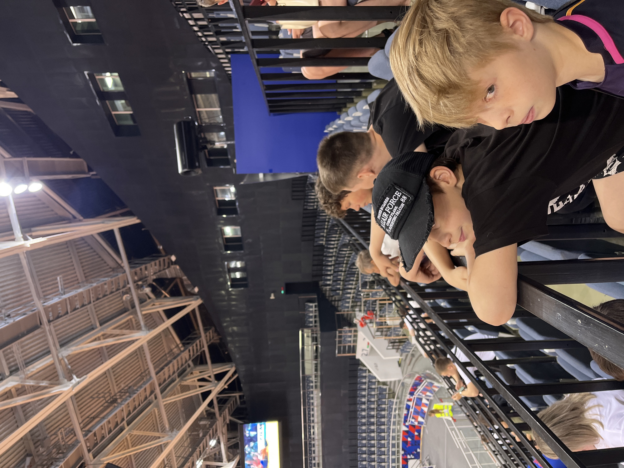In the foreground, two young boys are seated in the bleachers of an indoor arena. The boy on the right, with blond hair and blue eyes, is looking to his left. He is wearing a dark shirt with purple and pink trim. To his left, another boy with brown hair is wearing a black t-shirt and a black baseball cap. The cap has a patch that reads "FIGHTER BOMBER AIR FORCE COMBAT PLANE FIGHTING FALCON 88". The boys are leaning on a black metal railing, behind which are rows of empty blue seats.
In the background, the arena's interior is visible. The ceiling structure consists of metal beams and corrugated panels. The walls are primarily black, with some windows and a large blue screen or panel. The upper tiers of the arena seating are also visible, mostly empty, with some lights illuminating the space. To the left, a large screen displays an image of a sporting event. Other spectators are sparsely seated in the lower tiers. The overall environment suggests an indoor sporting event or concert venue. The location is identified as the city of Krasnoyarsk, Russia.