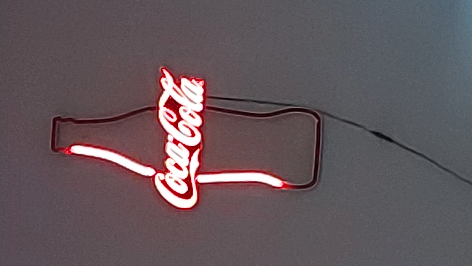 A neon sign is affixed to a light gray surface. The sign depicts a stylized Coca-Cola bottle with the words "Coca-Cola" rendered in bright white neon. The bottle shape is outlined in red neon. A black wire extends from the right side of the sign. The location is unknown.