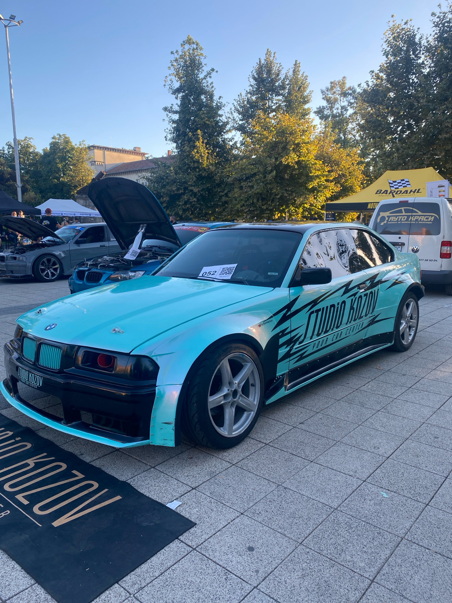 The image depicts a car show event in Pleven, Bulgaria. The foreground is dominated by a custom-modified turquoise BMW E36 coupe with black racing stripes and "STUDIO KOZOV YOUR CAR CENTER" branding. The car has a black roof, aggressive body kit, and aftermarket wheels. A black mat with "STUDIO KOZOV" branding is placed in front of the car. In the background, other cars with open hoods are visible, along with tents and banners. One tent displays "BARDAHL" branding, and a white van with "AUTO KRASИ" (meaning "Car Paints" in Bulgarian) is also present. The event appears to be taking place outdoors on a paved area, with trees and buildings in the background under a clear sky. The lighting suggests it is either early morning or late afternoon.