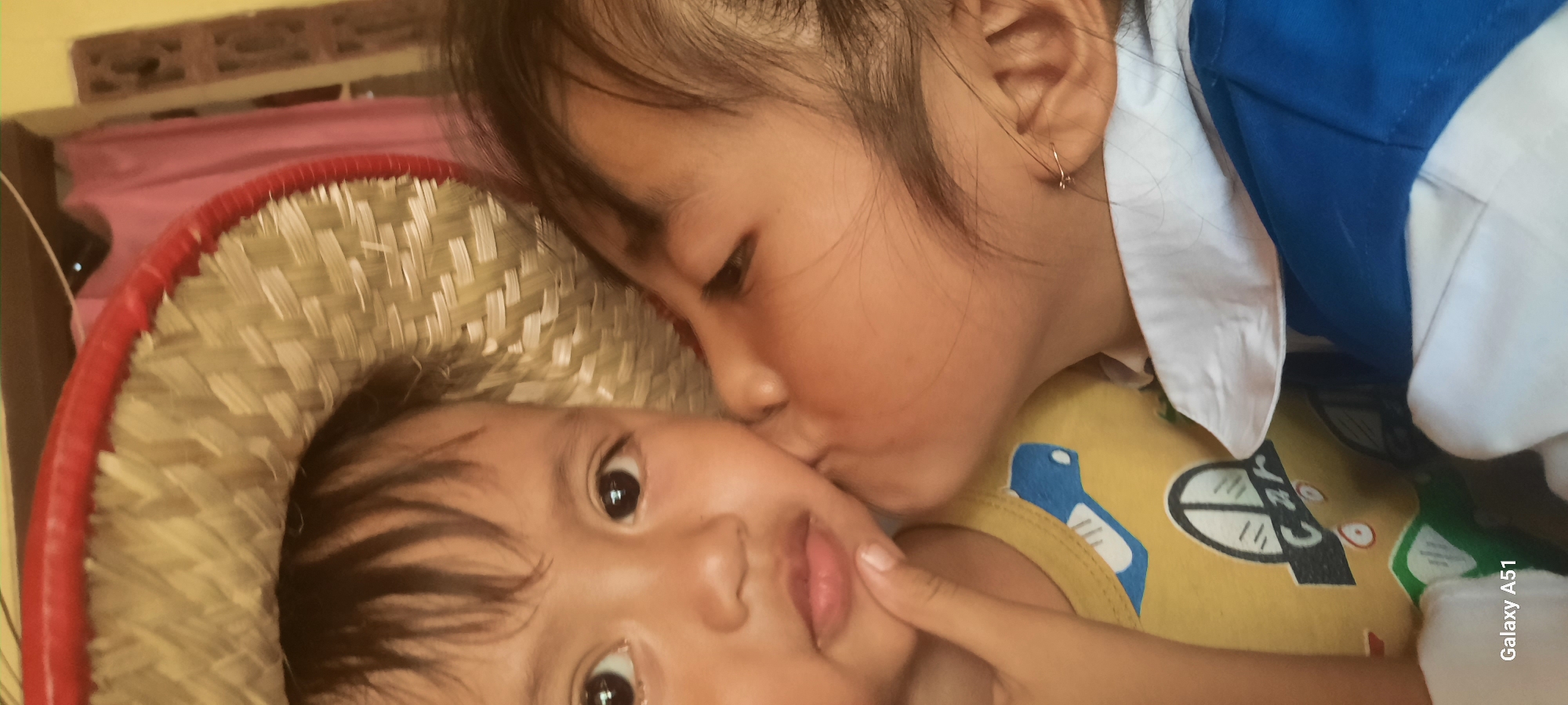 The image shows a close-up of two young children, likely siblings, in what appears to be an indoor setting. The child in the foreground, who is younger, is looking directly at the camera with wide, dark eyes. They are wearing a yellow shirt with a cartoon print. The older child is leaning in and kissing the younger child on the cheek, their eyes closed in a tender gesture. The older child is wearing a blue and white uniform, possibly a school uniform, with a white collar.
Both children have dark hair. The younger child appears to be nestled in a woven straw hat with a red trim, suggesting a possible outdoor or cultural element to their environment. The background is slightly out of focus but shows hints of what could be a wall with patterned bricks and a pink fabric. The lighting suggests it is daytime. The image evokes a sense of warmth, affection, and familial love. The text "Galaxy A51" is visible in the lower right corner, indicating the device used to capture the photograph. The location context provided is Jatitengah, Indonesia.