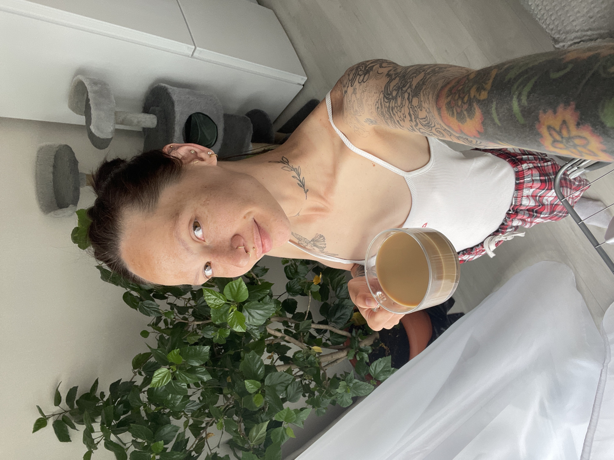 A selfie shows a woman with a nose ring and multiple ear piercings, holding a cup of coffee, standing indoors. She has tattoos on her chest and arm. The woman is wearing a white tank top and red and white plaid pants. A large green leafy plant is in the foreground, and a cat tree is partially visible in the background, along with white cabinets. A white curtain is present to the right side of the frame. The location is not discernible from the image.