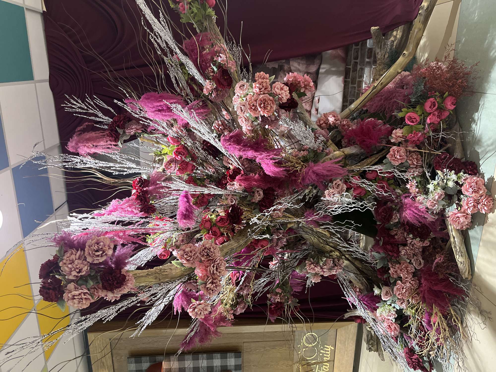 The image depicts a large, elaborate floral arrangement in what appears to be an indoor event space. The arrangement is predominantly composed of flowers in shades of pink, red, and burgundy, interspersed with feathery pink plumes and silvery, branch-like accents. The floral structure is built around a framework of large, weathered pieces of wood.
In the background, a dark red velvet curtain is draped. Above the floral arrangement, a section of the ceiling is visible, featuring a grid of yellow, blue, and green tiles. To the left of the arrangement, a wooden partition with a plaid pattern is partially visible, along with a golden sign that reads "ANCO Family." The flooring is a light-colored tile or polished concrete. The lighting suggests an interior setting.
No specific location in the city of Veliko Tarnovo, Bulgaria is identifiable from the visual elements in the image.