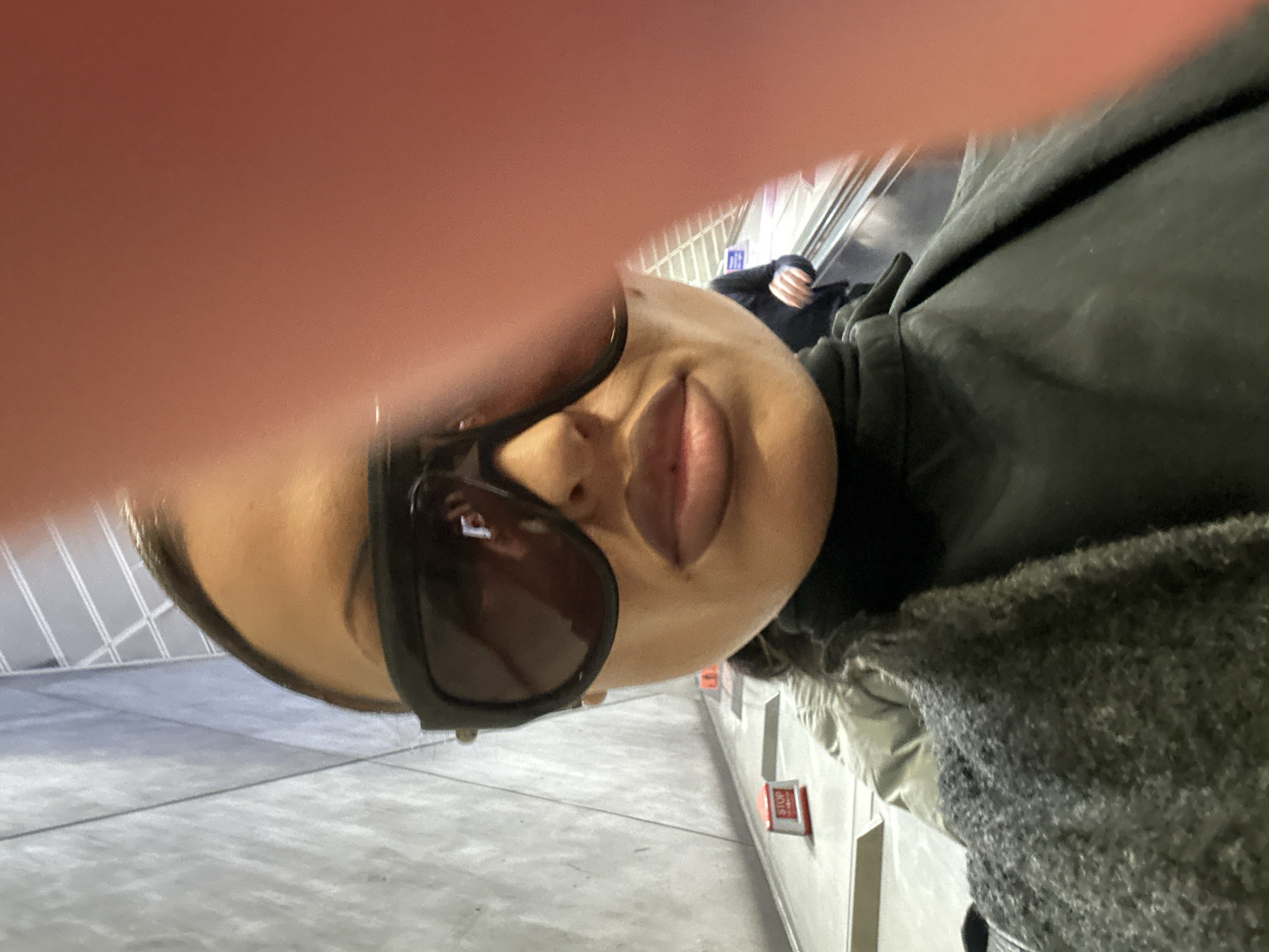 A selfie-style photograph shows a person wearing sunglasses and a jacket, likely on an escalator. The background reveals a modern setting with metallic elements and linear patterns suggestive of architecture. A "STOP" sign can be seen, and there appears to be another person on the escalator behind the main subject. The location is identifiable as a London Underground station in the United Kingdom, specifically Canary Wharf station on the Jubilee line, recognizable by the distinctive silver and white exterior features.