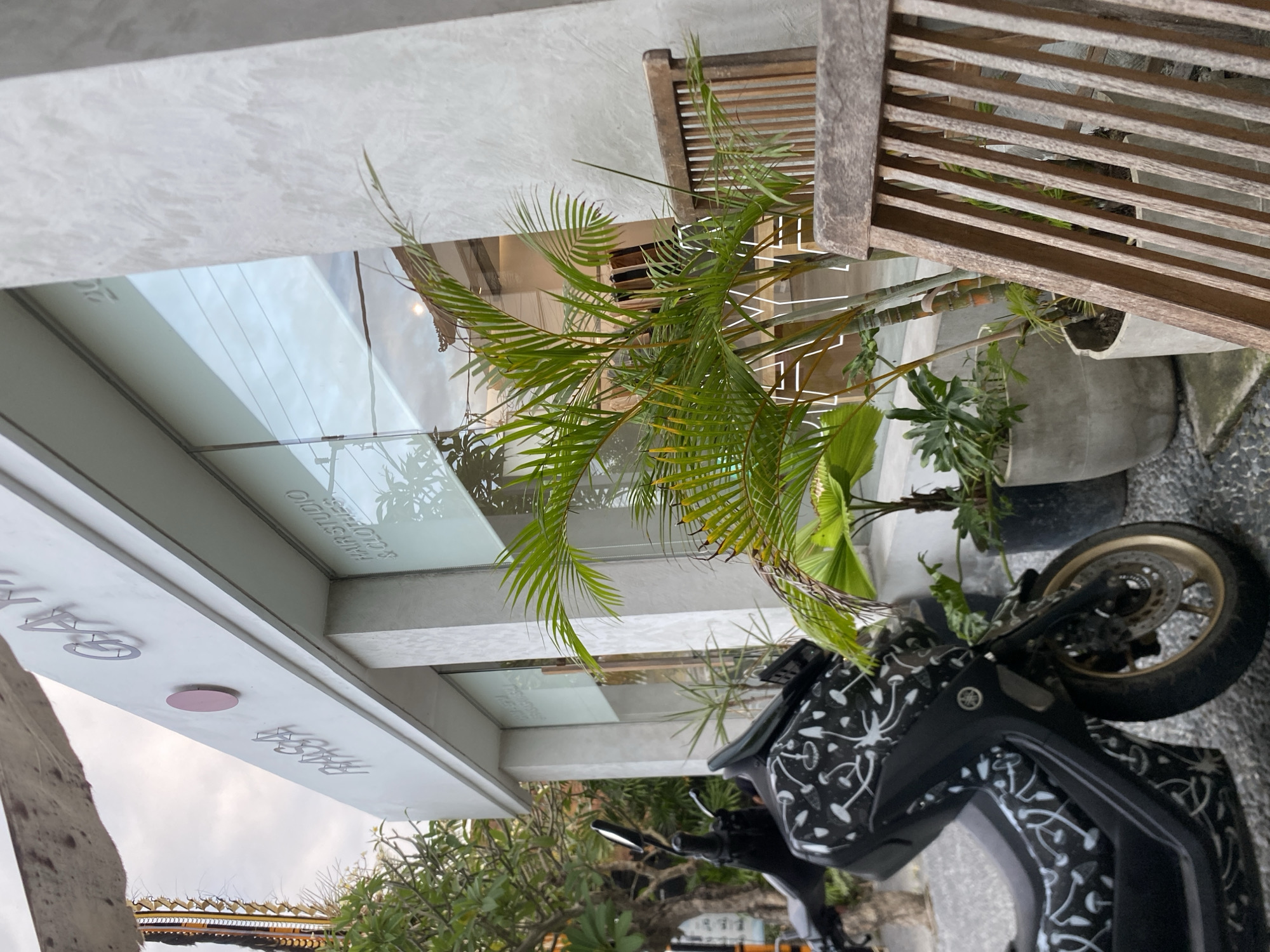 The image shows the exterior of a modern building with signage reading "RASA" and "GA". The building appears to be a hair studio and clothing store. In front of the building, there is a black motorcycle with a white mushroom pattern. To the right of the motorcycle, there are several potted plants, including a palm tree. A wooden chair is visible in the foreground. The setting is likely an outdoor commercial area in Mengwi, Indonesia. The sky is overcast.