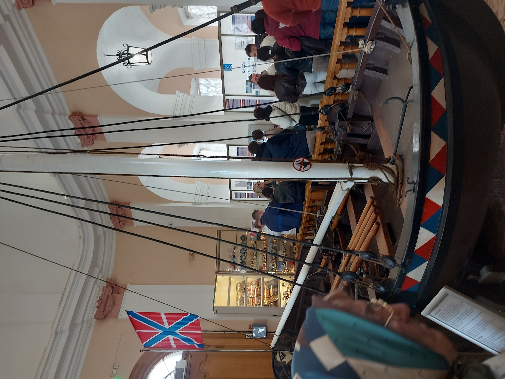 A low-angle interior shot features a ship, potentially a replica, moored in what appears to be a historical building. People are lined up along the deck, looking towards the forward area of the ship. A Russian naval ensign, specifically the St. Andrew's flag, hangs from the ship's mast. In the background, there's a souvenir display case suggesting this may be a museum. Considering the St. Andrew's flag, and the general museum-like setting, this is likely in the Central Naval Museum, located in Saint Petersburg, Russia. The building's architecture is ornate, with decorative moldings on the ceiling and walls.