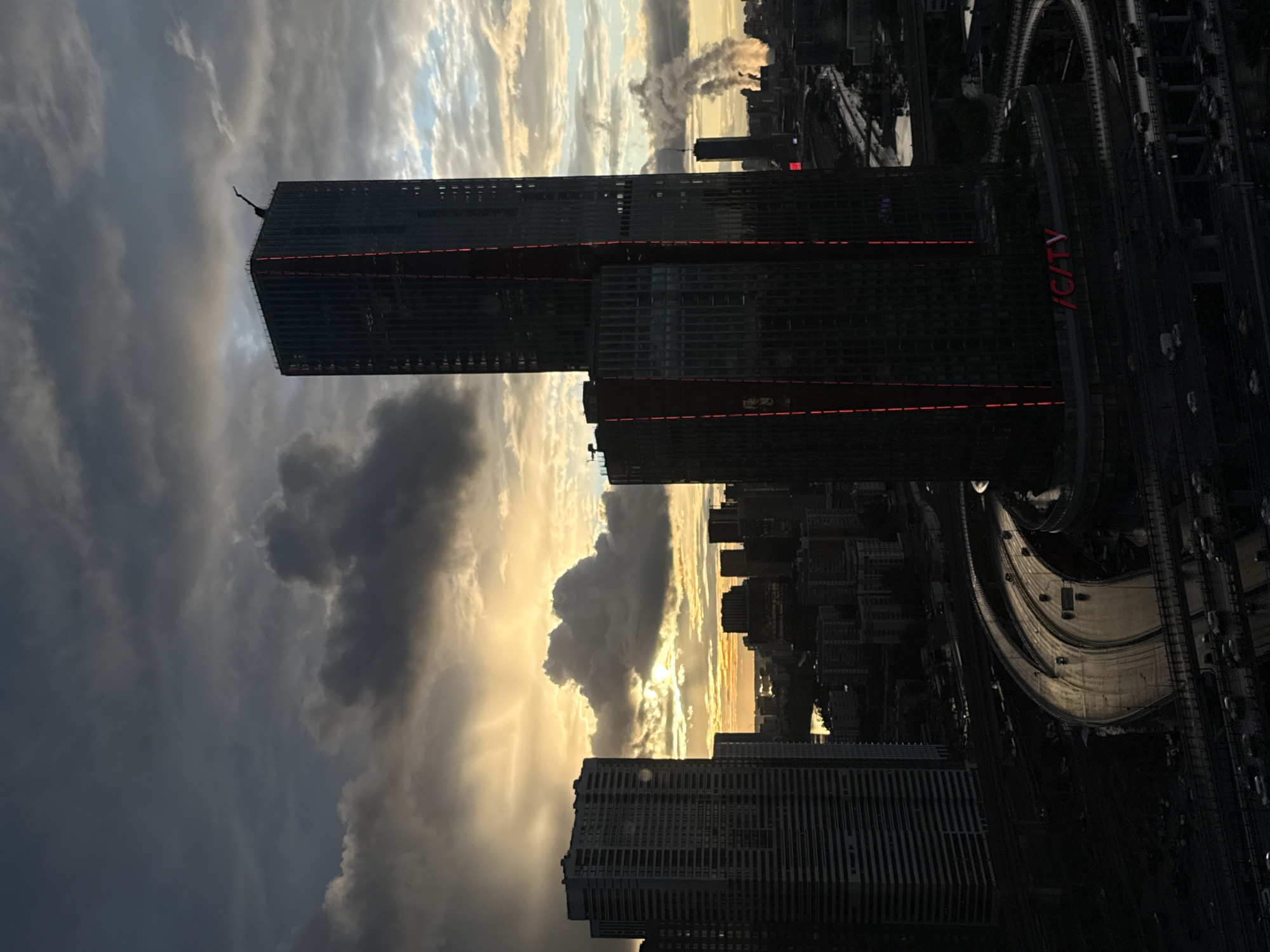 The image depicts a cityscape at dusk or dawn, possibly in Khimki, Russia, with tall buildings and infrastructure. A prominent skyscraper with red lights is visible on the right, and a large cloud formation illuminated by the sun dominates the left side of the frame. The roads around the skyscraper are visible with traffic.