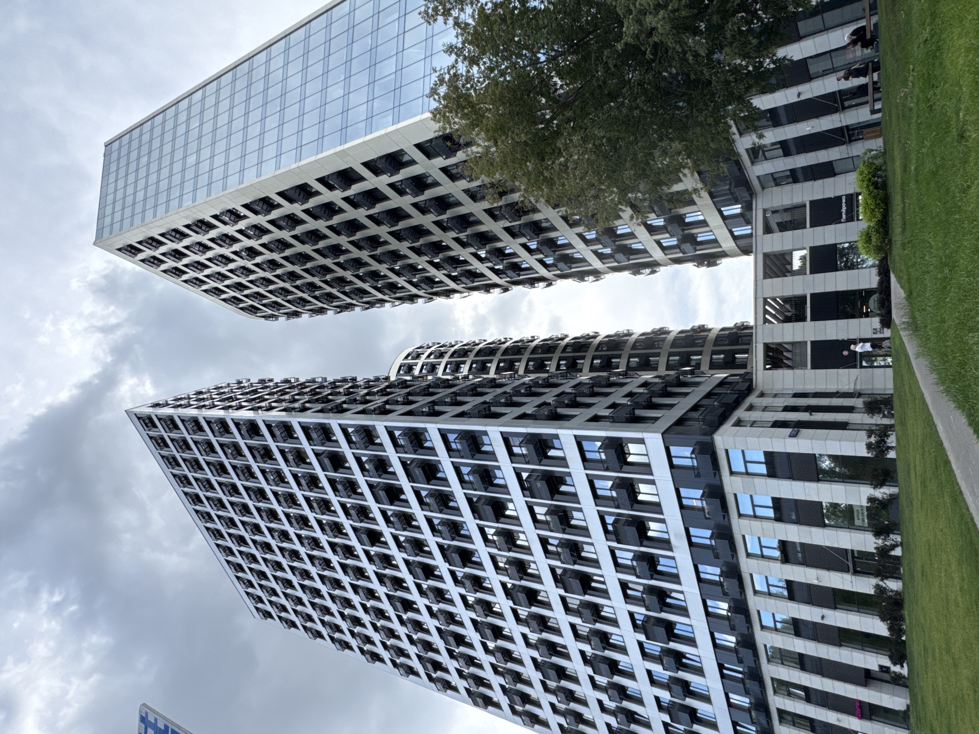 This image captures a low-angle view of several modern, high-rise buildings under a cloudy sky. The buildings feature repeating geometric patterns on their facades, with numerous windows and protruding balconies or modular units. One building on the right is predominantly glass, reflecting the sky. In the foreground, a neatly manicured green lawn slopes upwards, with a paved walkway cutting through it. Several people are visible in the distance, some sitting on benches and others walking. A lush green tree is situated to the right of the frame, partially obscuring one of the buildings. The architectural style suggests a contemporary urban development, likely in a metropolitan area. The signage "Пятёрочка" (Pyaterochka) on one of the lower building levels identifies it as a Russian supermarket chain, confirming the location as Moscow, Russia.