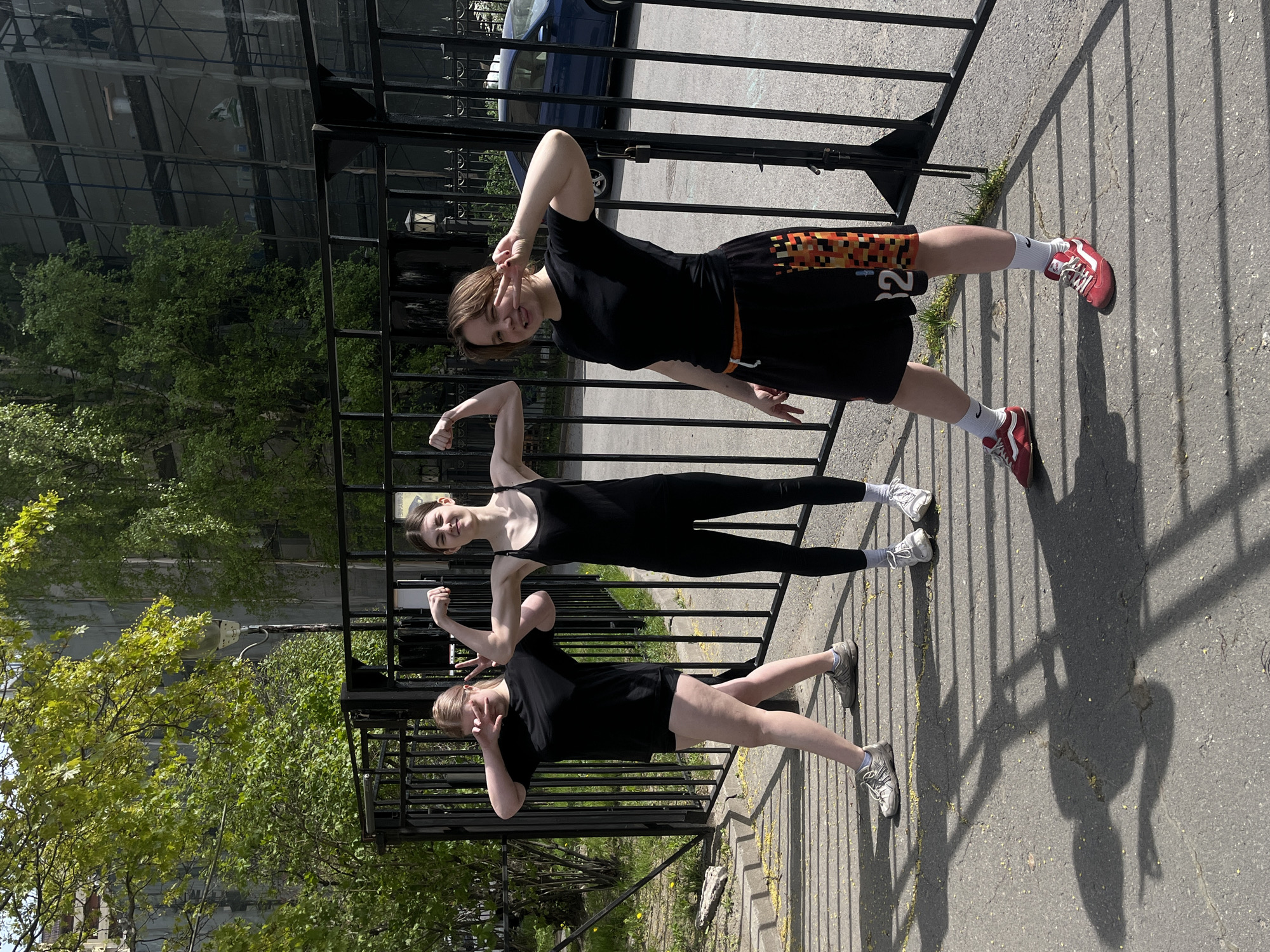 Three young women pose outdoors, positioned in a line against a black metal fence. Each woman wears athletic attire. The woman closest to the camera wears a black t-shirt and shorts, and is flashing a peace sign with her left hand. The woman in the middle wears a black leotard. The woman furthest from the camera wears a black t-shirt and black shorts with a colored pattern along the side. The background includes trees, parts of a building with reflective windows, and a blue car. The setting appears to be an urban area. The location is the city of Saint Petersburg, Russia.