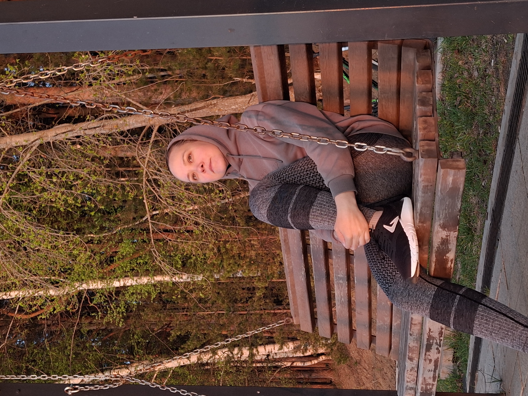 A woman sits on a wooden swing in what appears to be a park or wooded area. She is wearing a gray hoodie and patterned leggings, and she is holding onto the swing's support. The swing is suspended by chains, and behind the swing are trees with sparse leaves. Some grass is visible in the background. The location is in a city in Russia, which, in this case, is Chaikovsky.